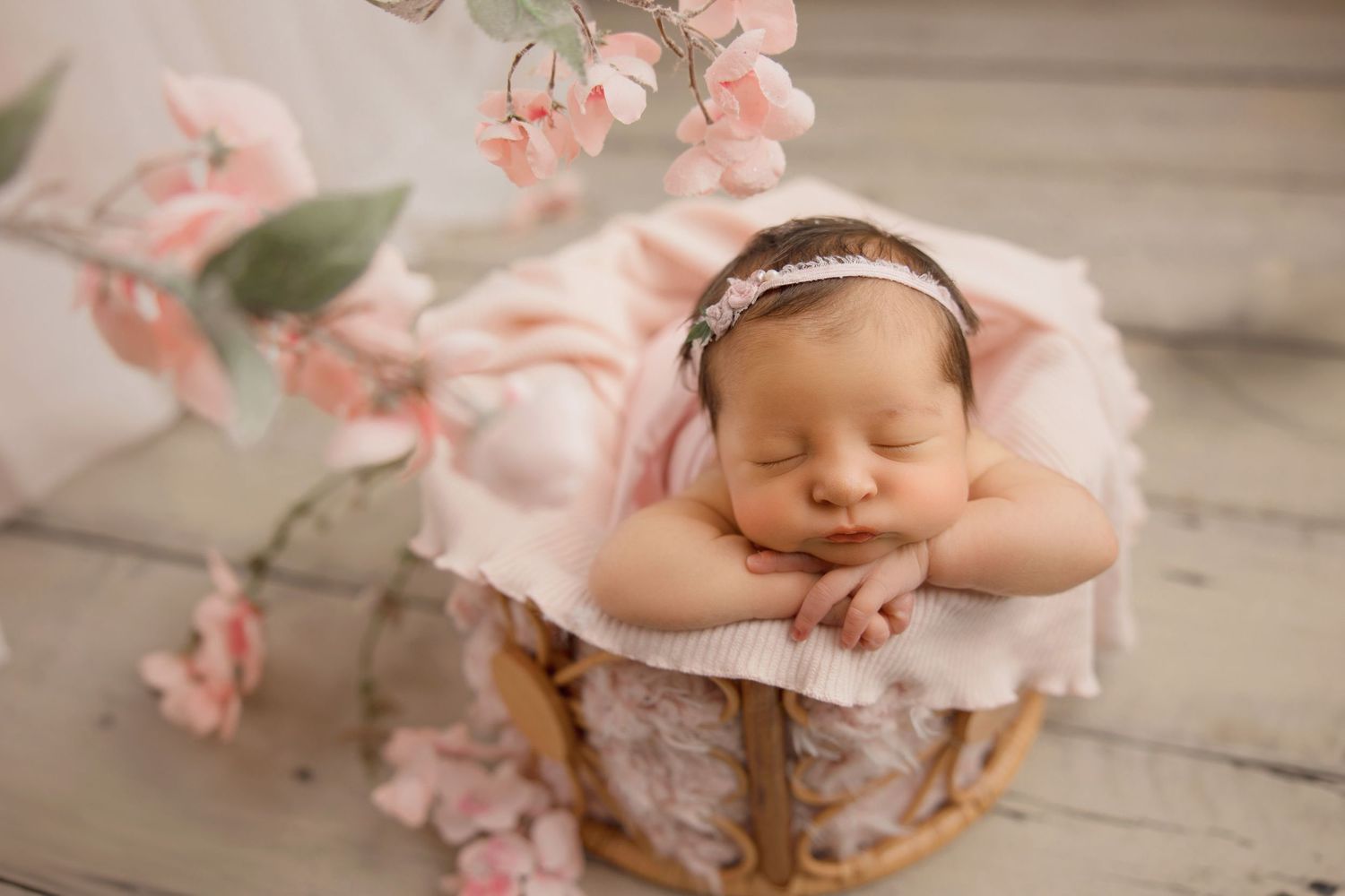 Sleeping newborn in pink outfit rests peacefully in wooden basket surrounded by delicate cherry blossoms.