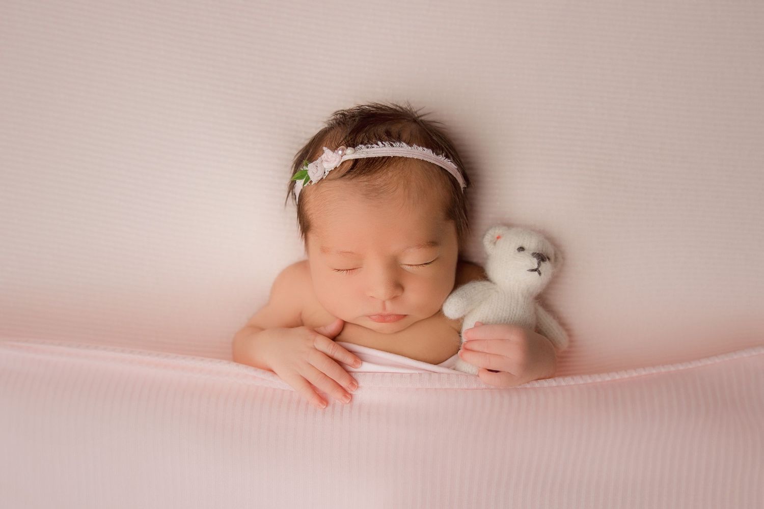 A sleeping newborn baby wearing a white headband lies peacefully on soft pink fabric in a professional photo setting.