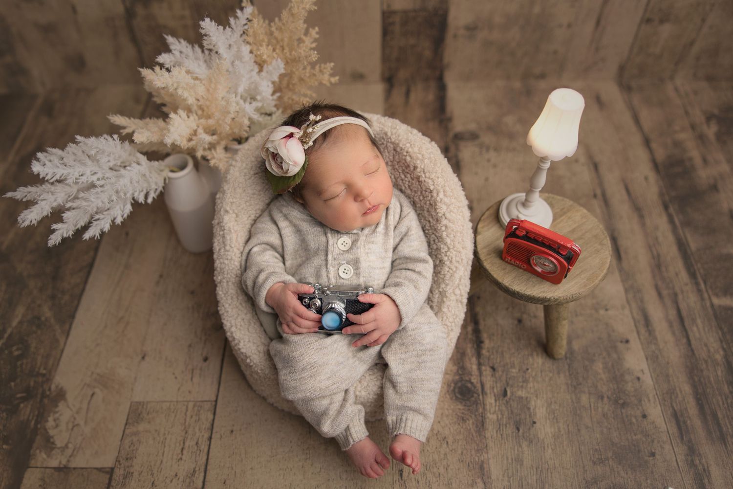 Sleeping newborn in gray outfit holds video game controller on wooden floor with white decor and lantern.