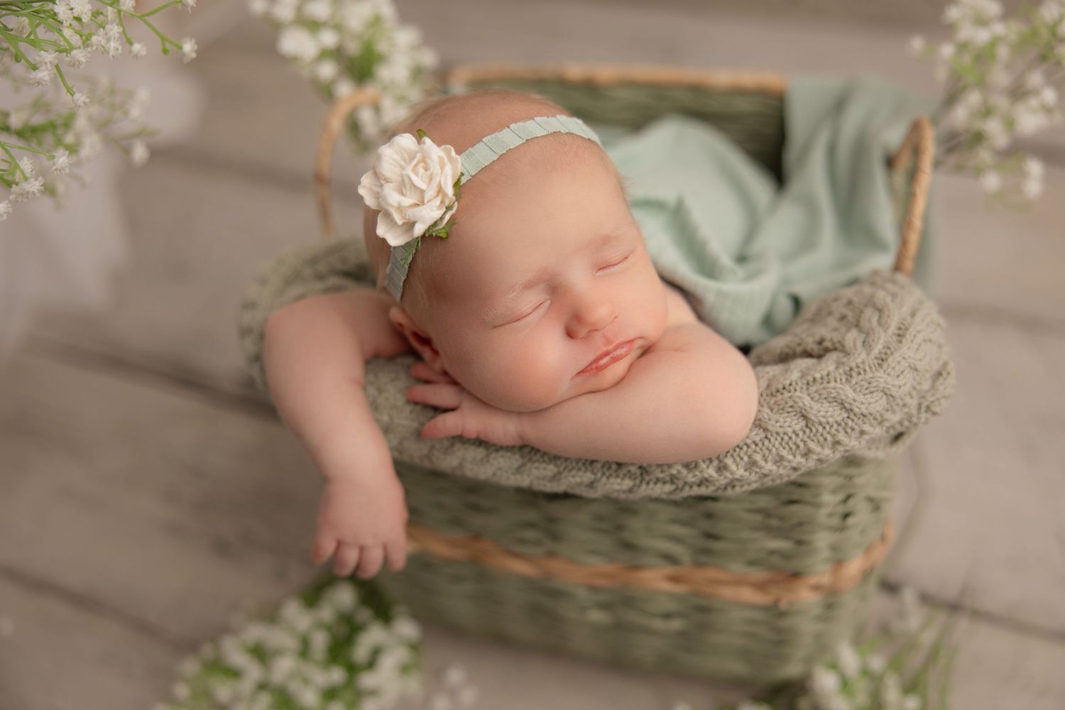 A sleeping newborn baby wrapped in soft mint green fabric rests peacefully in a woven basket during a professional photoshoot.