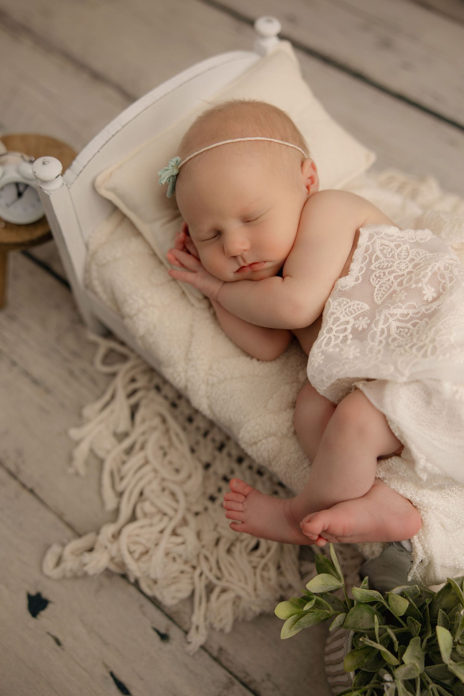 Sleeping baby in white lace on cozy cream blanket with decorative fringe and small plant on rustic wooden floor.