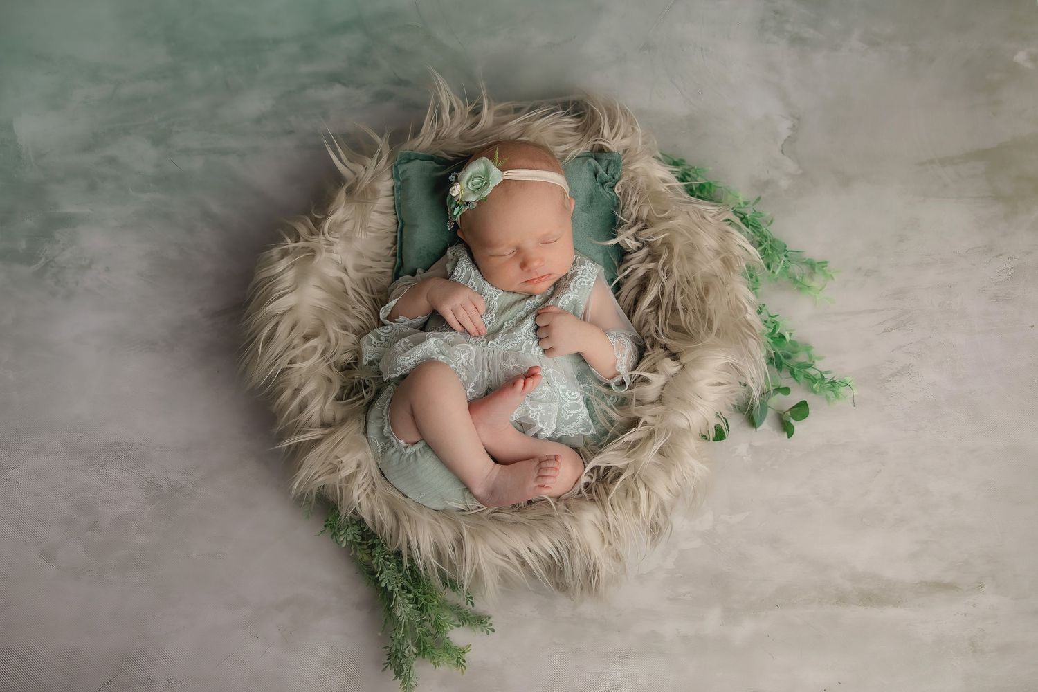 Sleeping newborn wearing mint green outfit lies on fluffy cream fur with decorative greenery accents in studio setting.