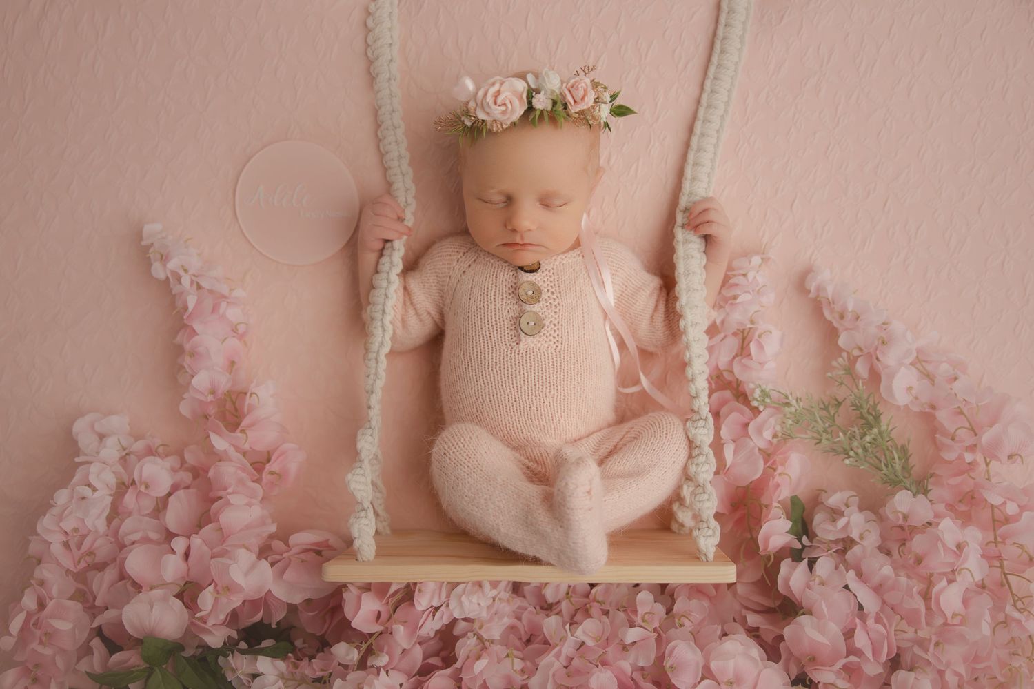 A sleeping baby on a wooden swing surrounded by soft pink cherry blossoms and wearing a floral crown.