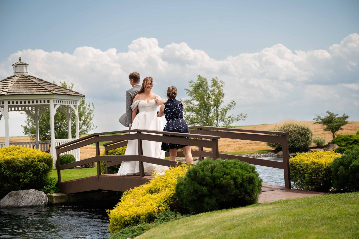 Wedding party stands on a decorative wooden bridge overlooking a landscaped garden with white gazebo and pond.