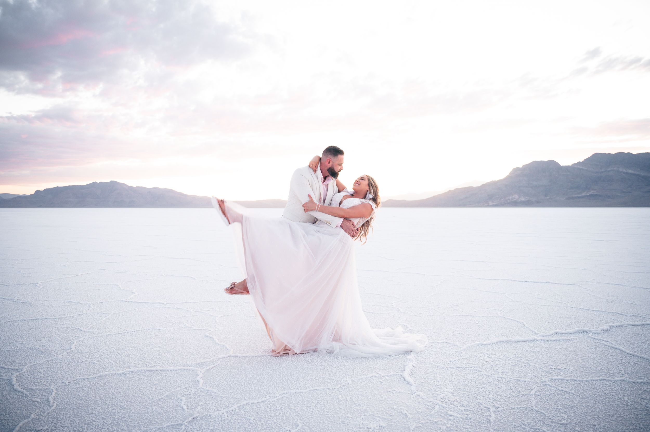 A couple in wedding attire dances on the salt flats with mountains in the distance at sunset. Salt flats elopement wedding photographer