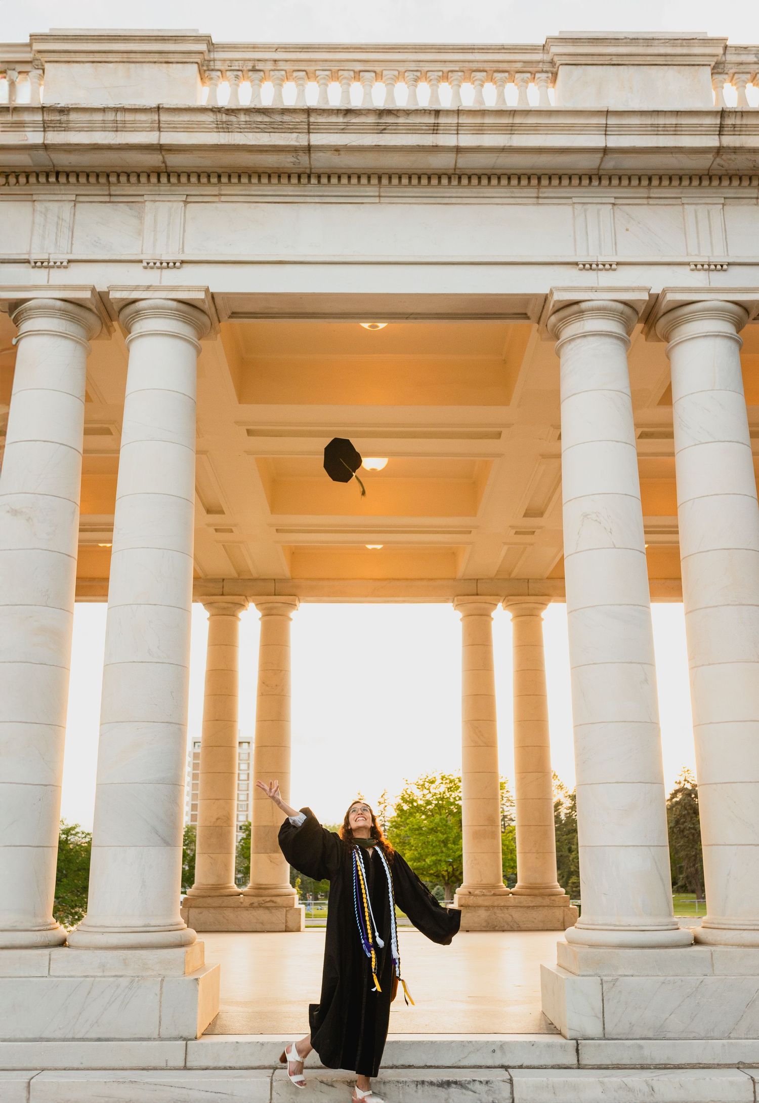 Colorado School of Mines graduate celebrating with cap toss under grand white columns of historic building during golden hour