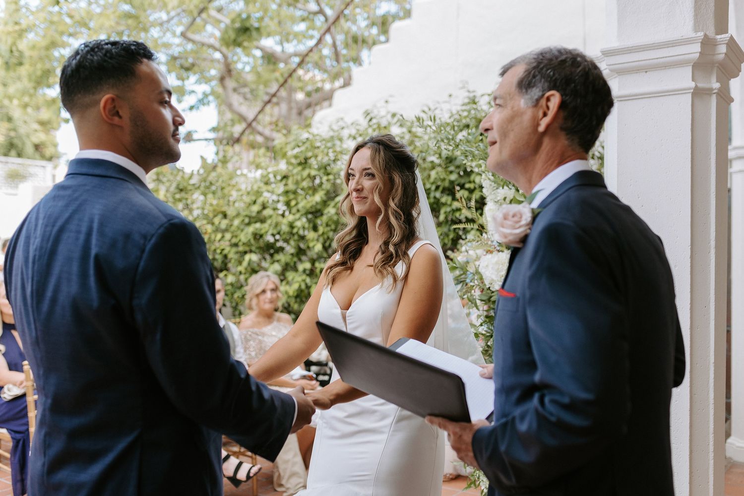 Couple exchanging vows at outdoor wedding ceremony with officiant holding book, bride in white gown, groom in navy suit at Darlington House.