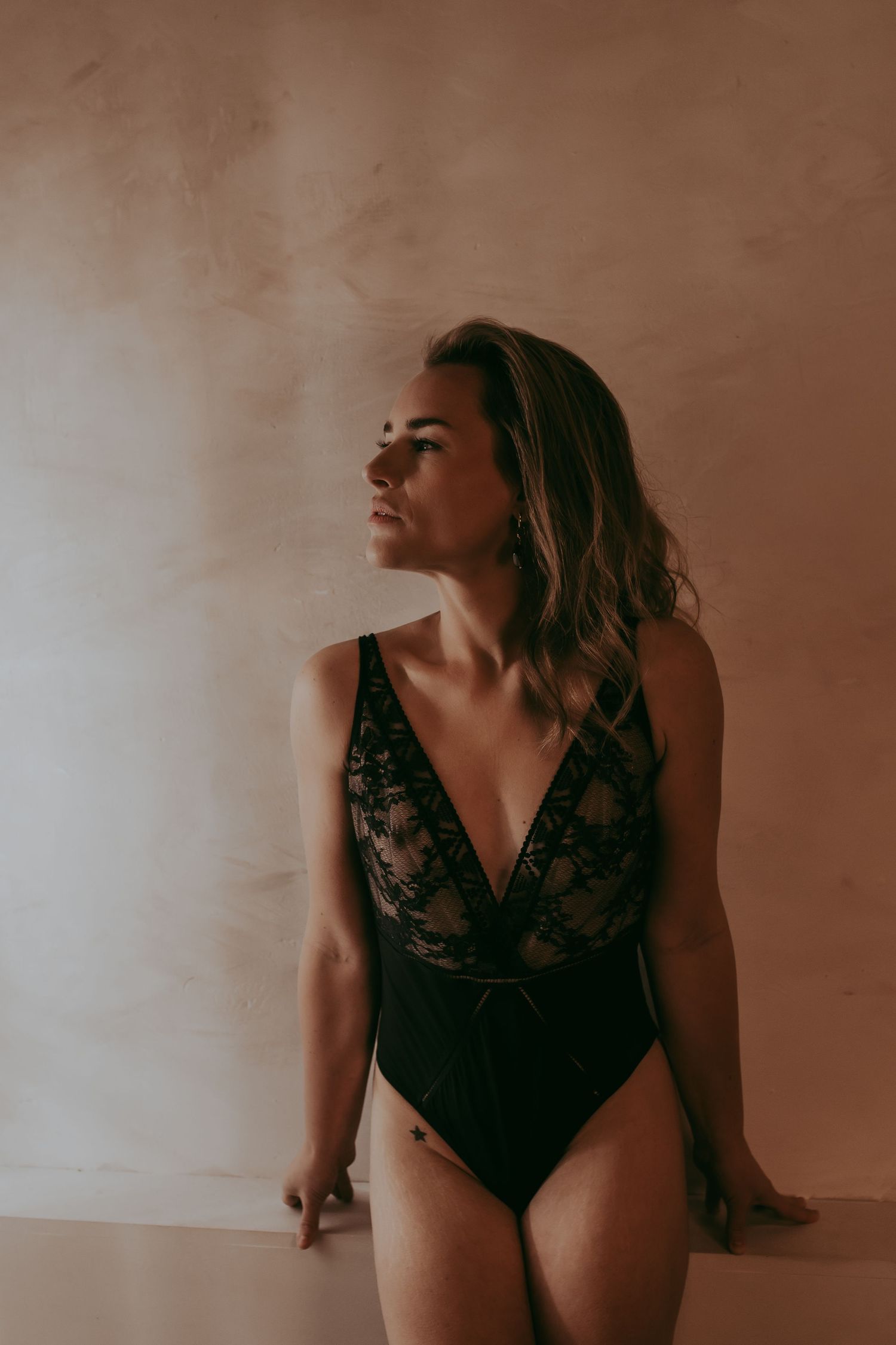 Vrouw in een kanten lingerie body met een donkere achtergrond half zittend.