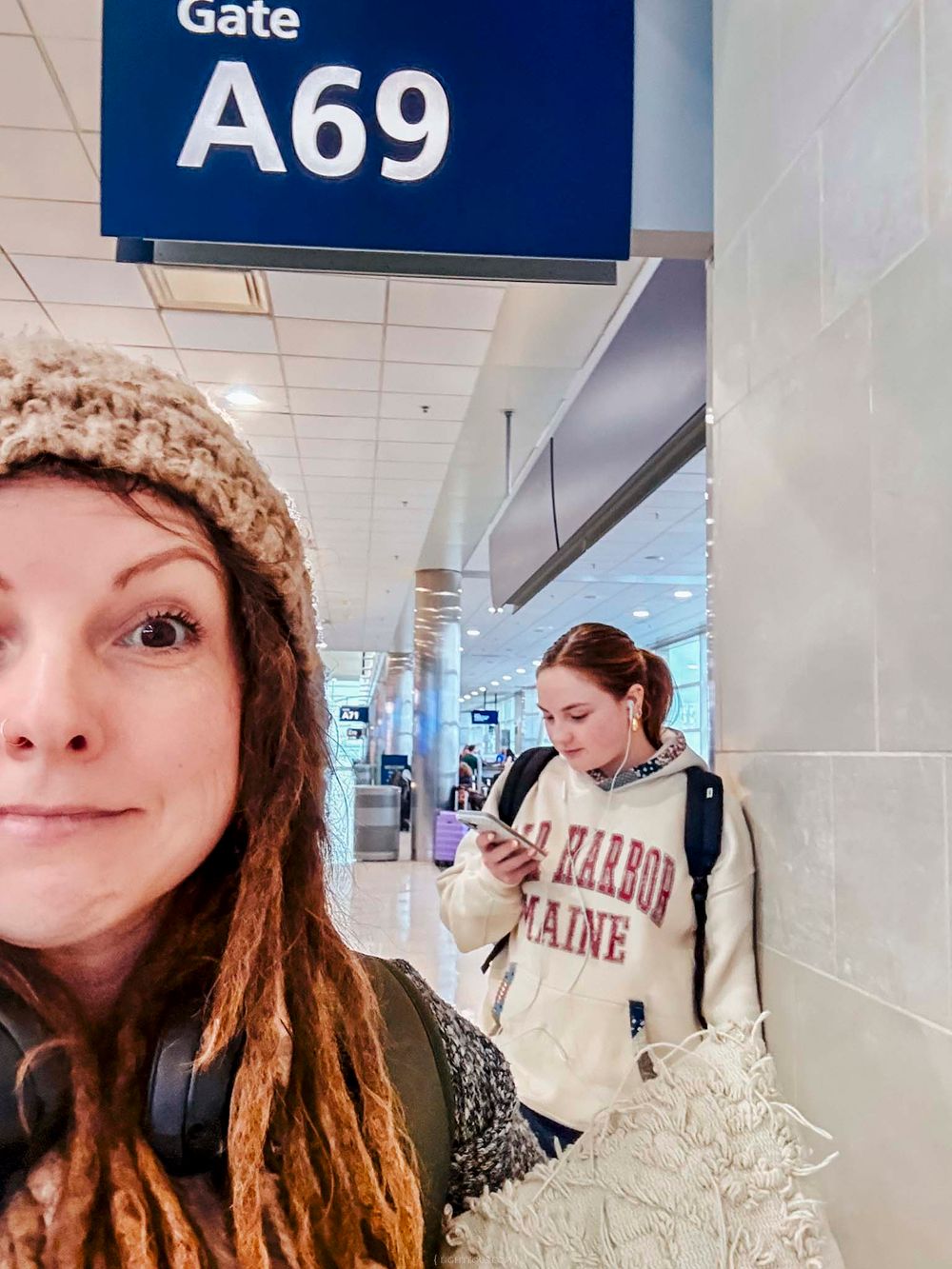 Woman taking selfie at airport Gate A69 with another woman in a Bar Harbor Maine sweatshirt visible behind her.