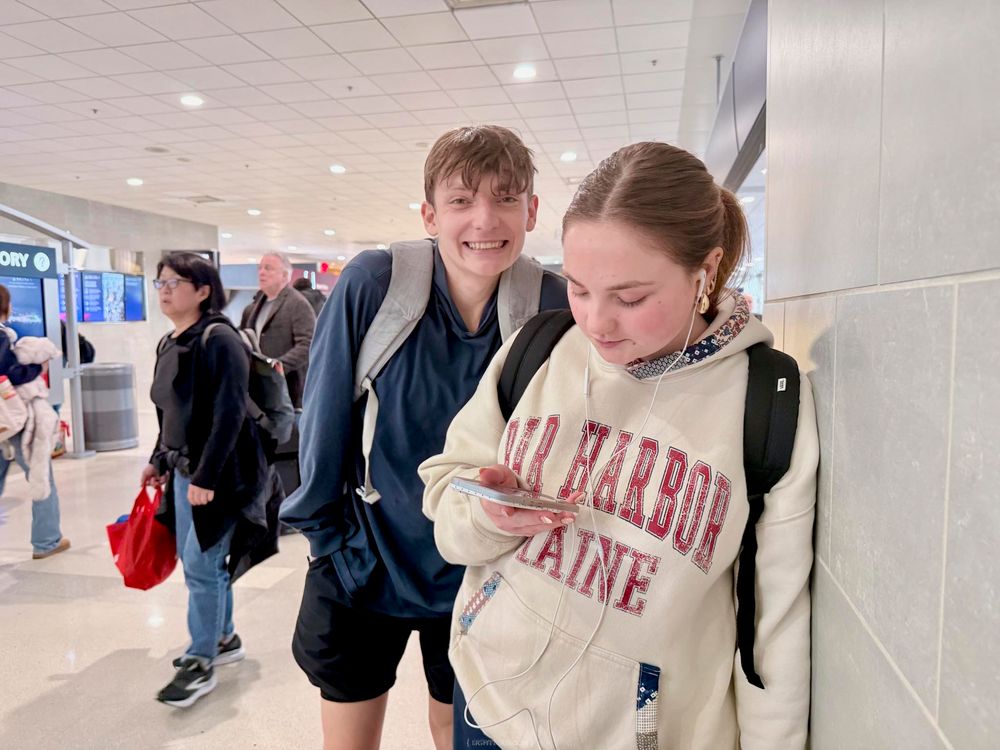 Two teenagers at an airport, boy smiling with backpack, girl looking at phone wearing Bar Harbor Maine hoodie.