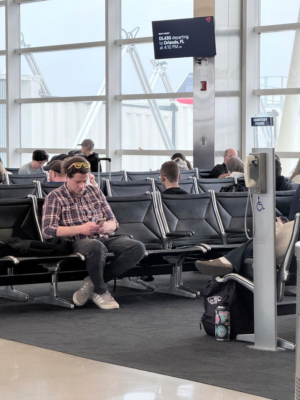 Man in plaid shirt uses phone at airport gate seating area with departure board visible overhead.