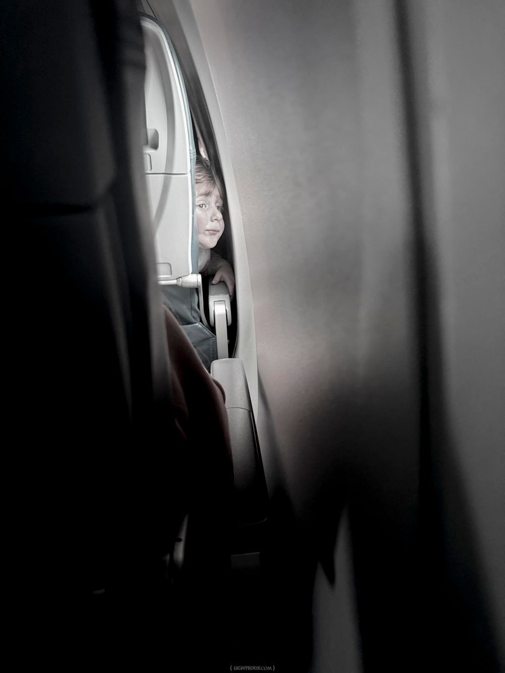 Child peering through airplane seat gap, partially visible face illuminated in dark cabin interior.