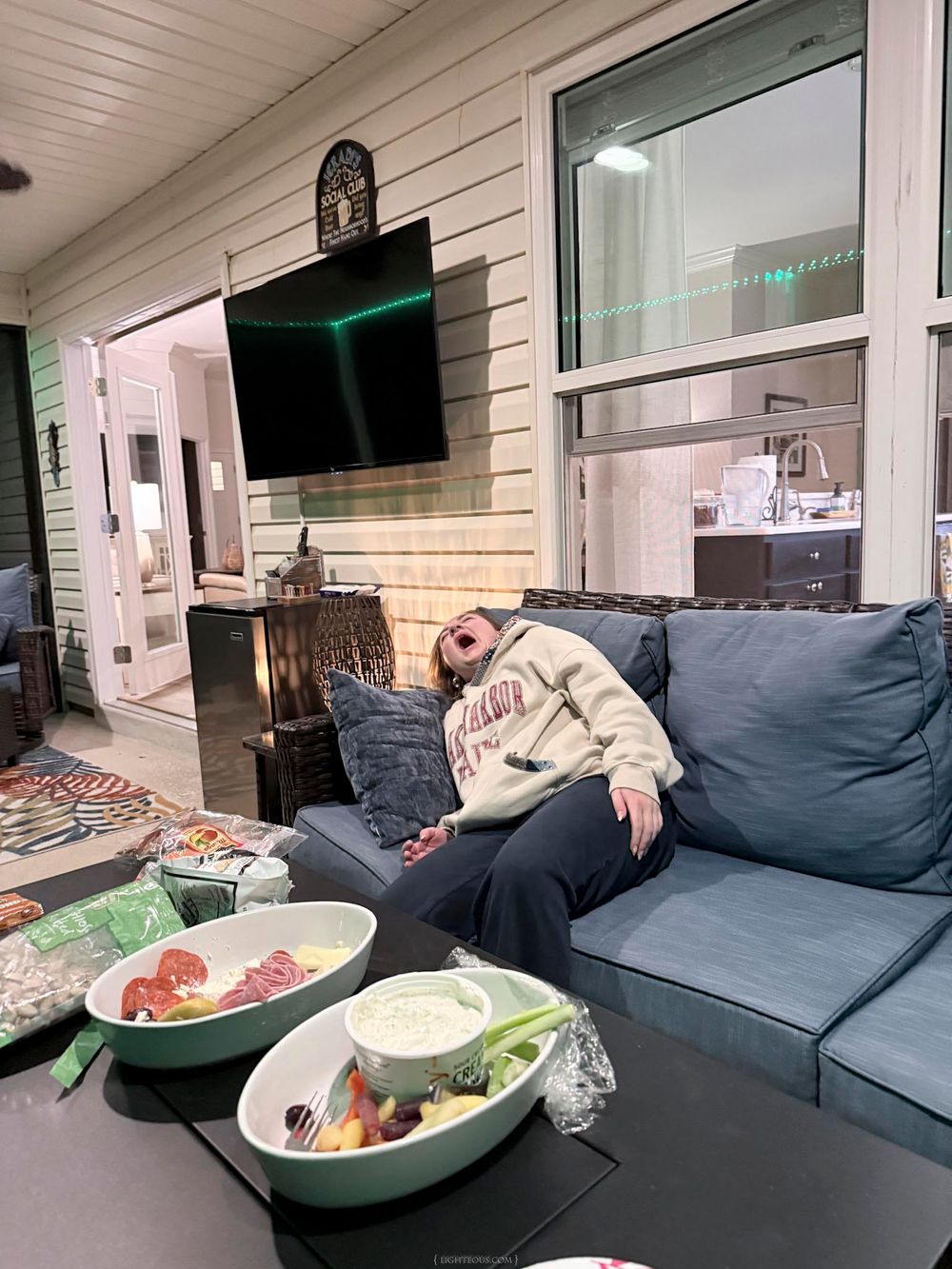Person relaxing on blue outdoor sofa near coffee table with food bowls, patio TV mounted on wall behind.