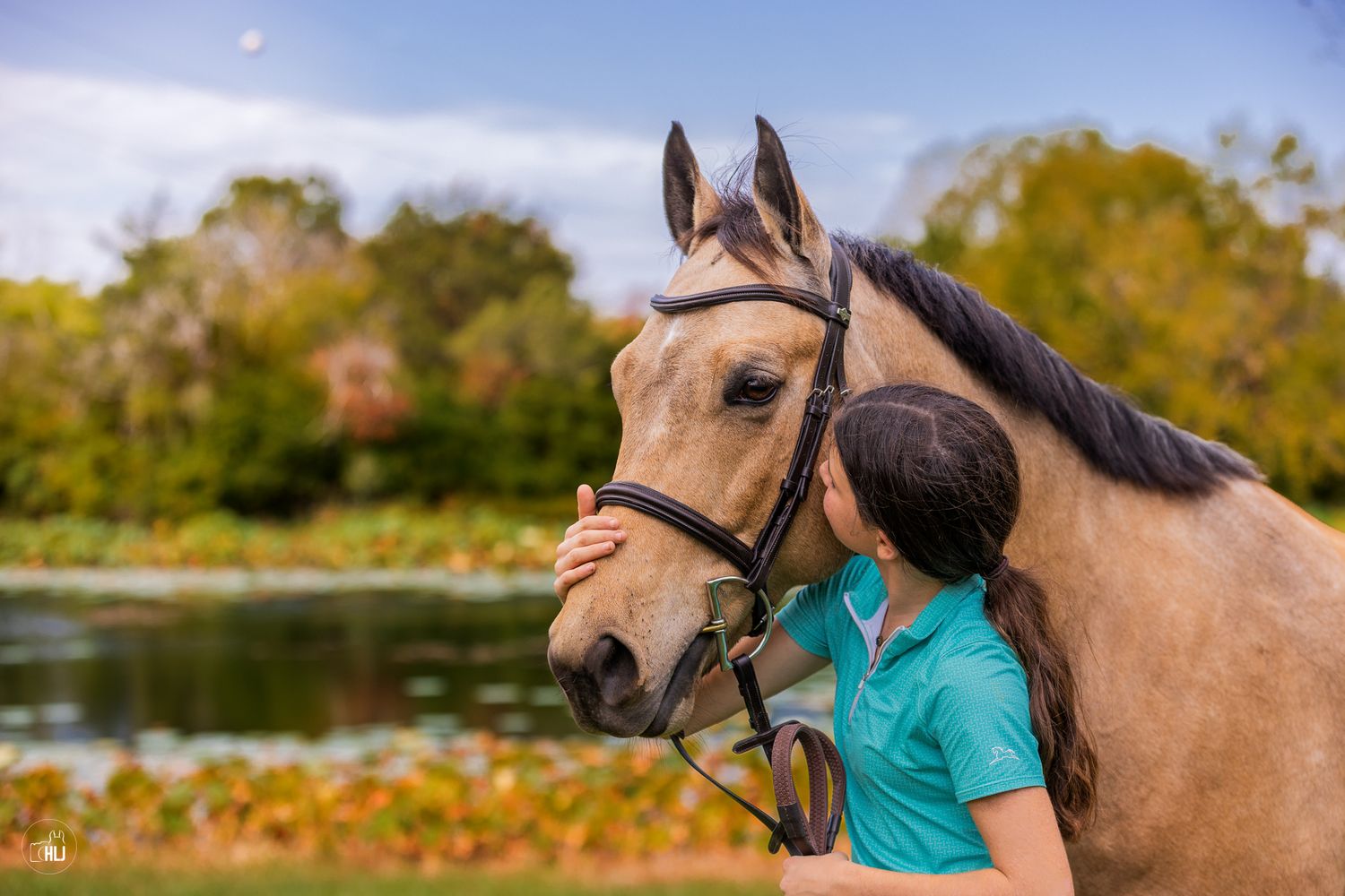 A young girl in a teal shirt kisses a buckskin horse wearing a bridle near a serene pond with autumn trees.