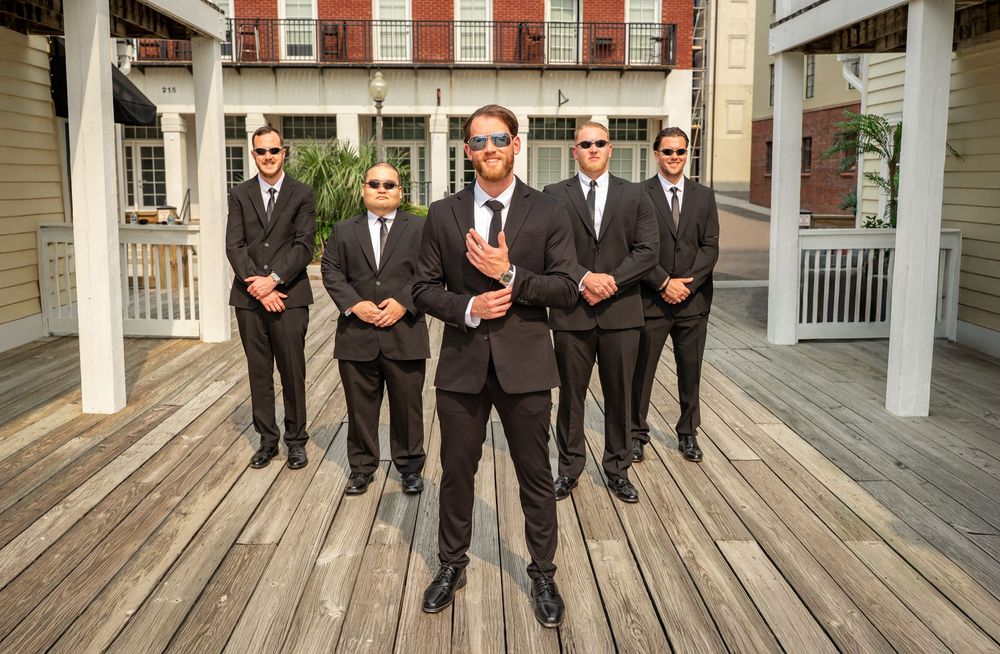 A group in formal black suits and ties walking in sequence along a wooden porch deck.