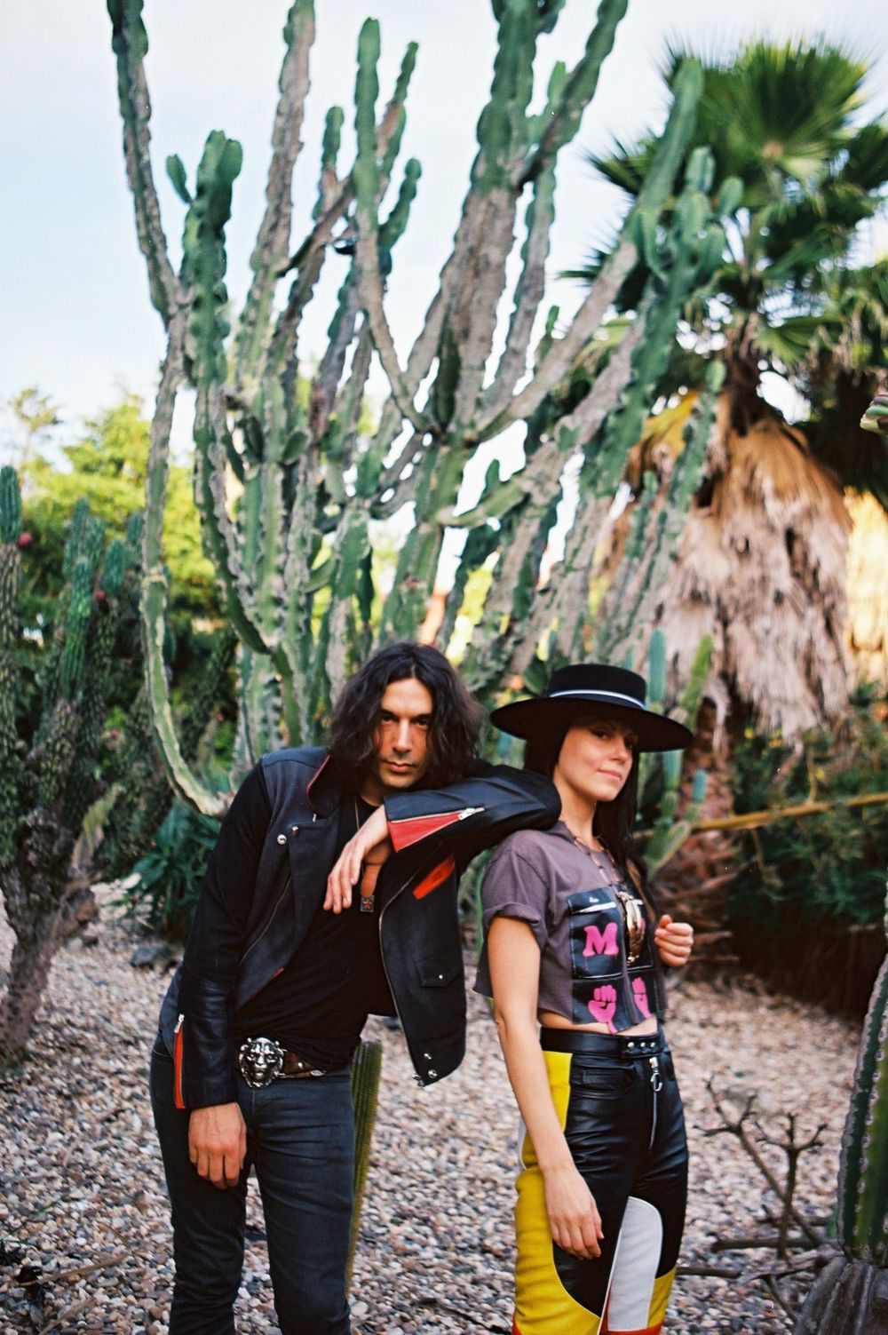 Two people pose together in a desert garden setting with tall cactus plants in the background.