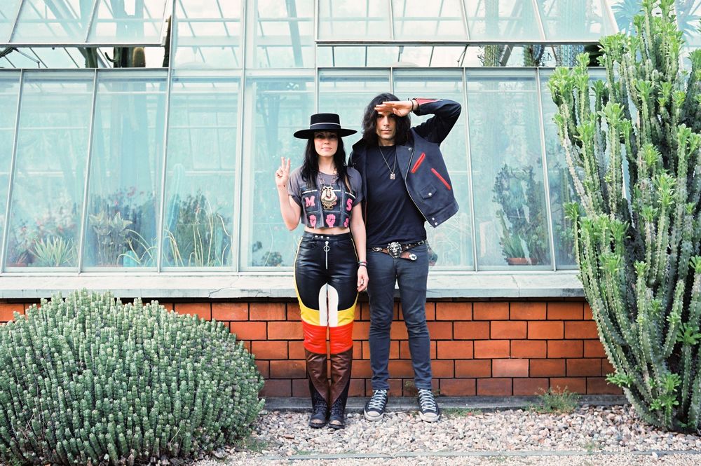 People stand near a greenhouse with brick walls and tall cactus plants growing along the exterior glass panels.