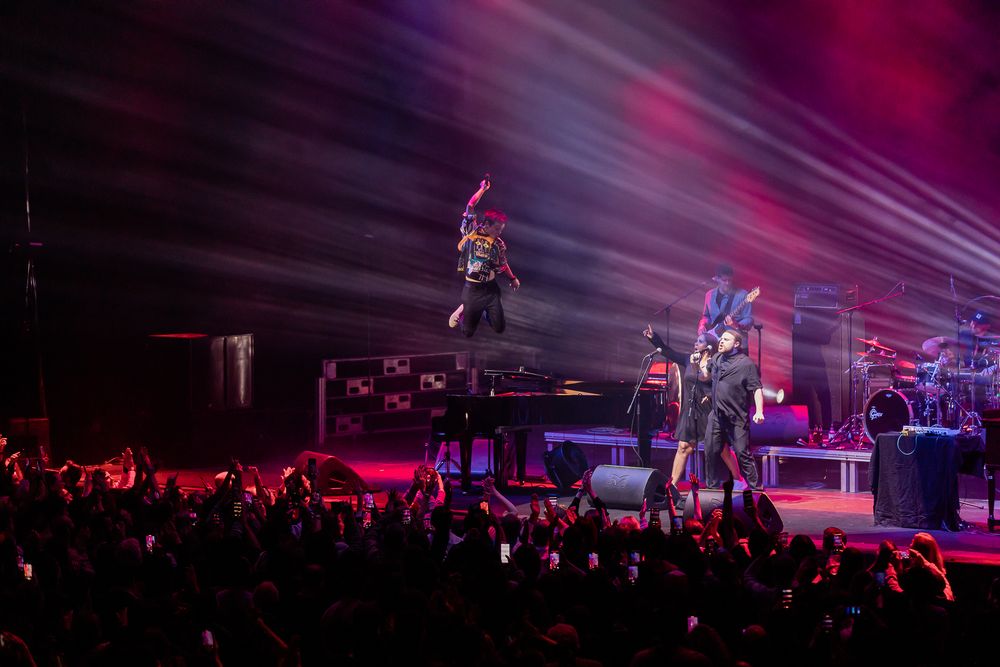 Musicians perform on stage with dramatic purple lighting and beams while a crowd watches at a live concert venue.