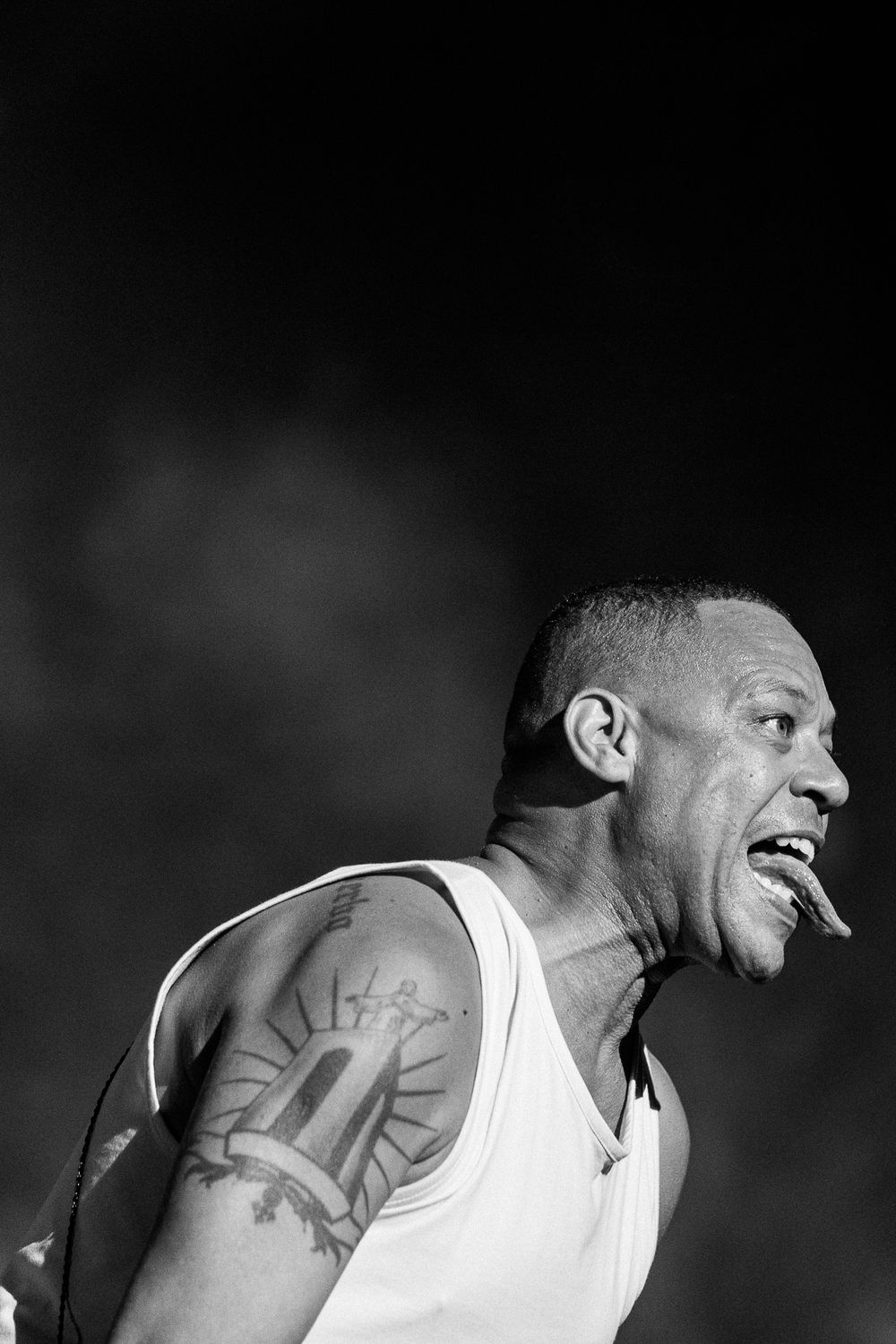 Black and white photo of a performer in a white tank top with visible arm tattoos singing energetically on stage.