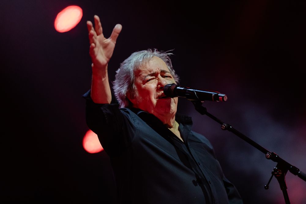 A performer sings into a microphone on stage with dramatic red lighting effects in the background.
