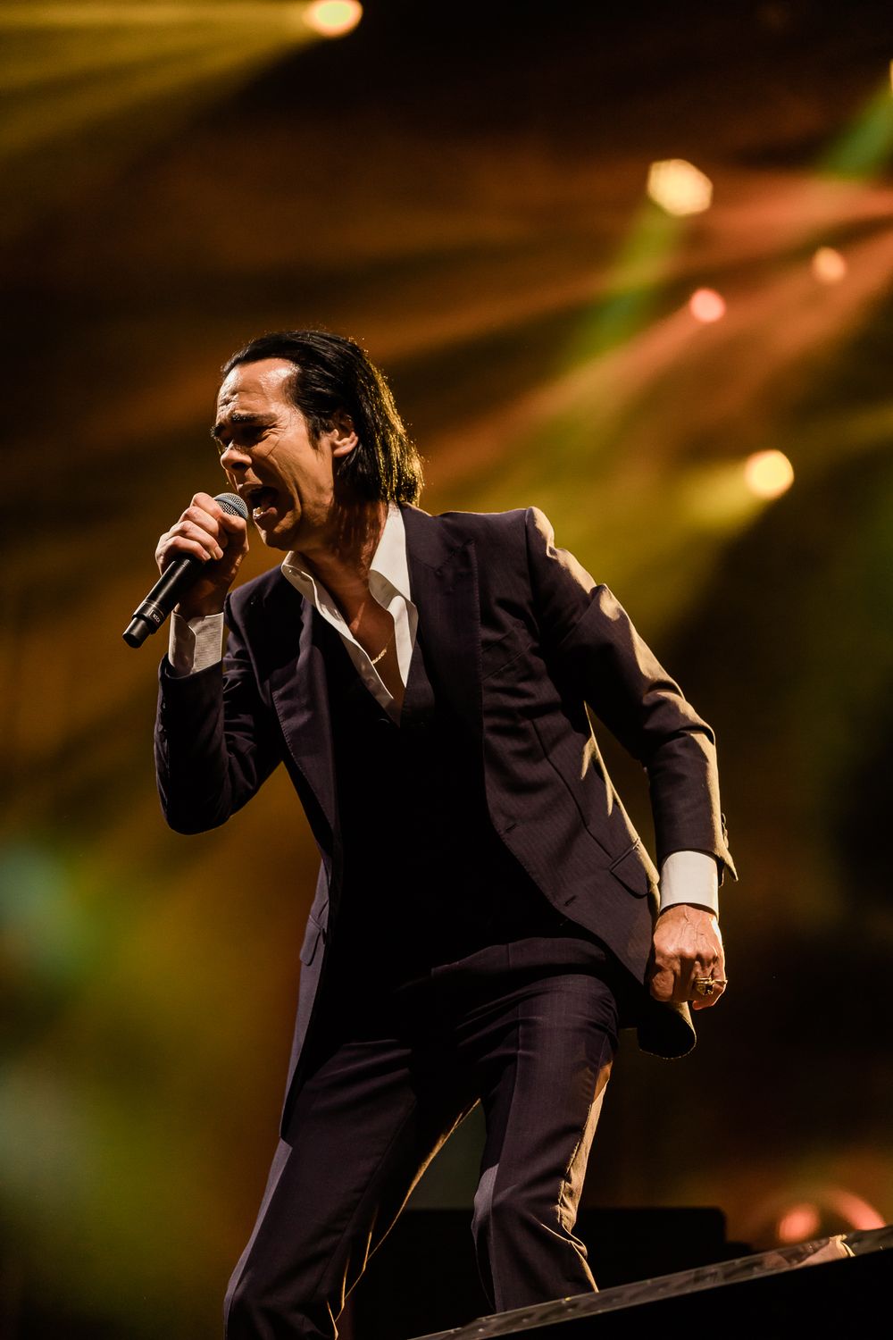 A performer in a dark suit sings passionately on stage under dramatic concert lighting.