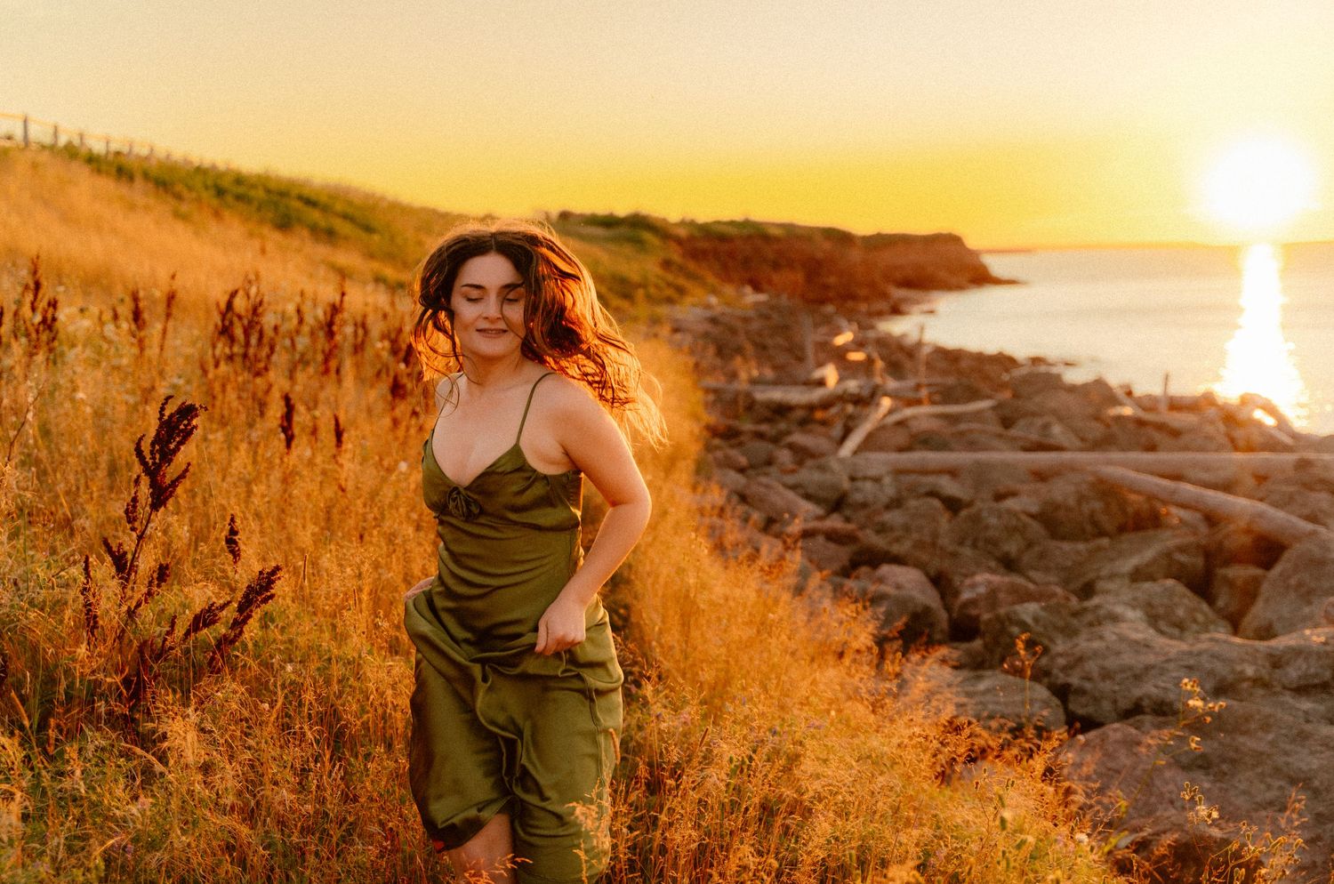 Figure in olive green outfit poses on coastal cliff at sunset.