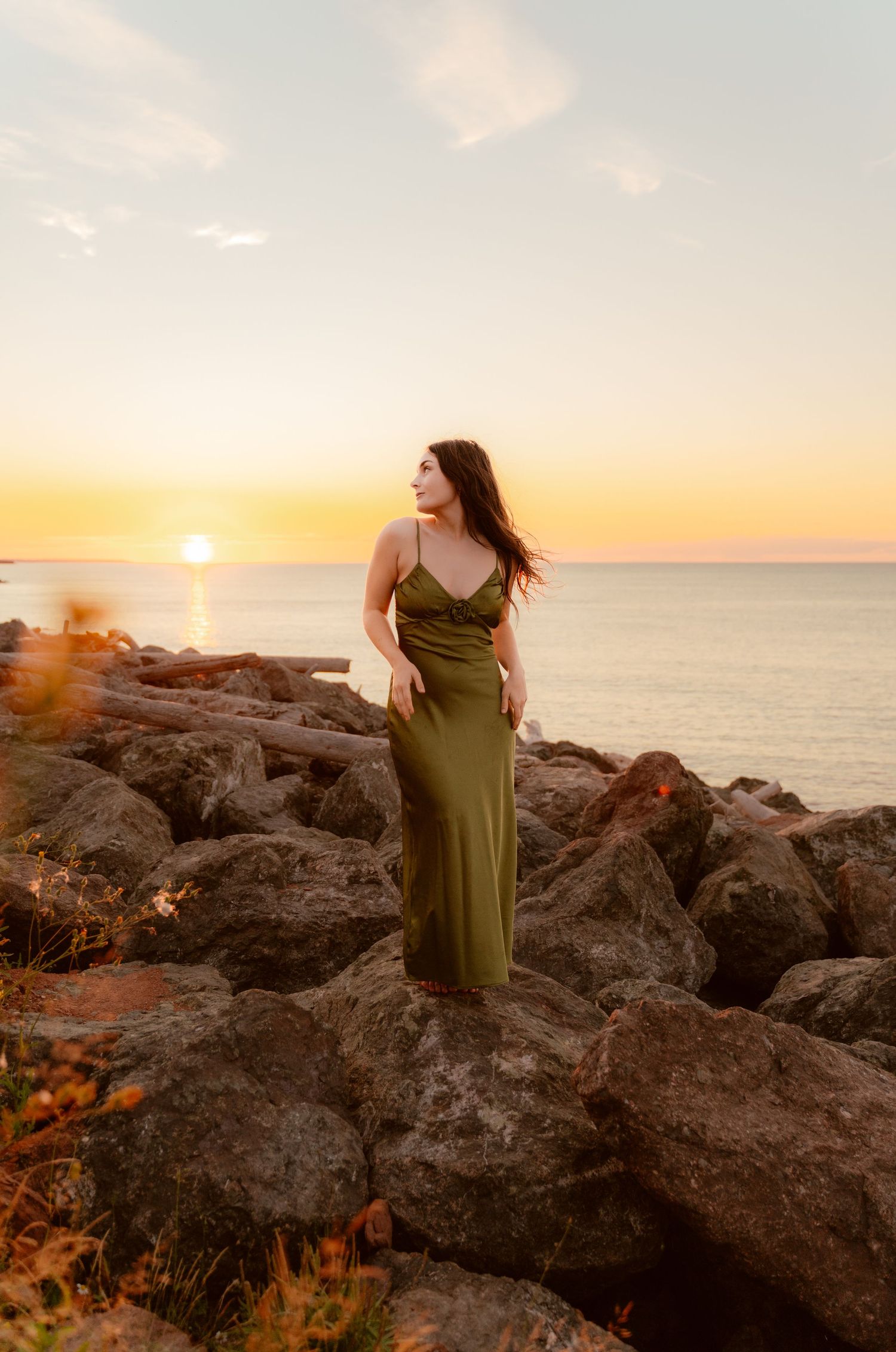 A figure in a green dress poses on rocky coastal cliffs during a golden sunset over the ocean.