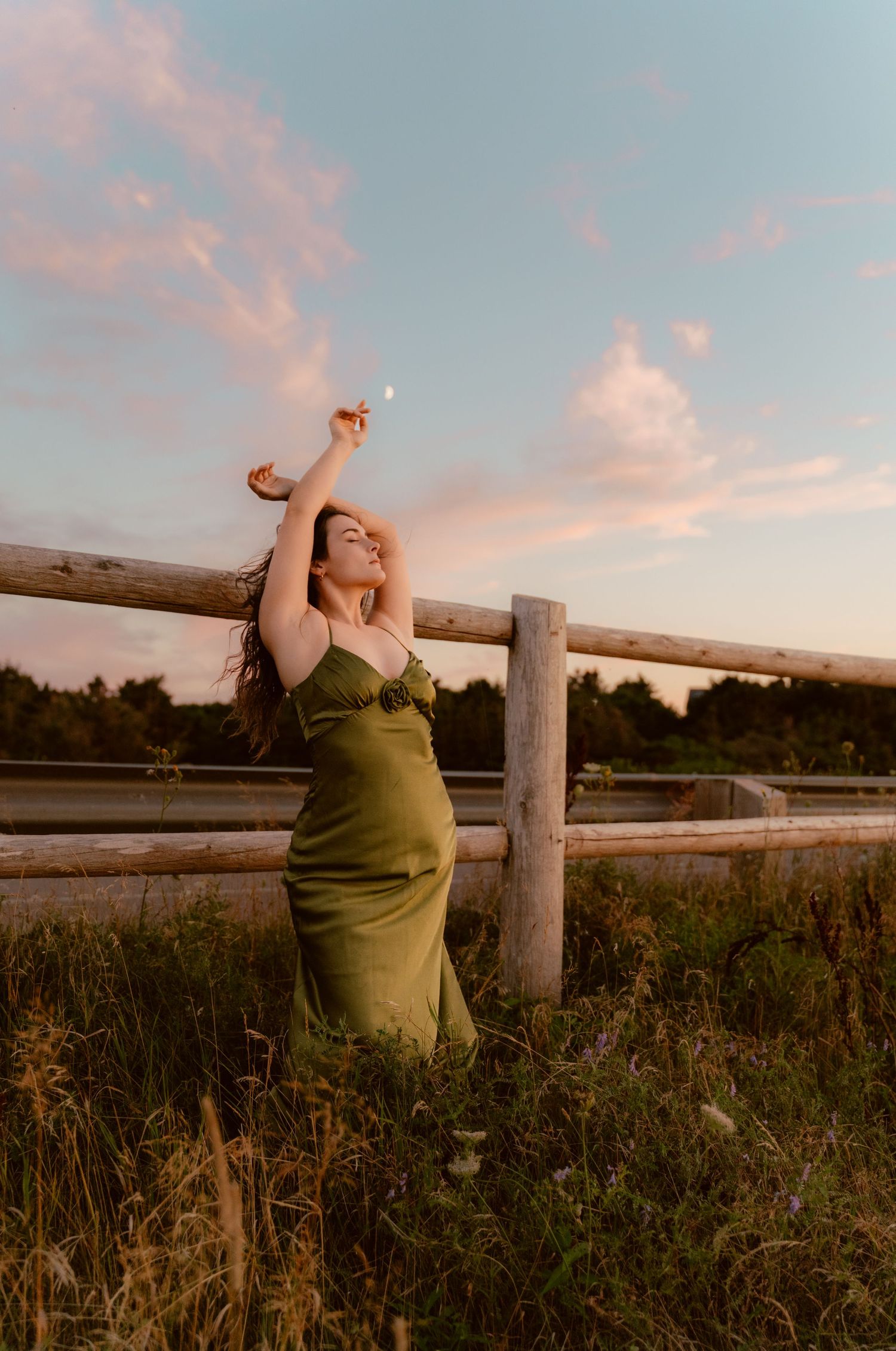 A person in an olive green dress poses dramatically against a wooden fence at sunset.