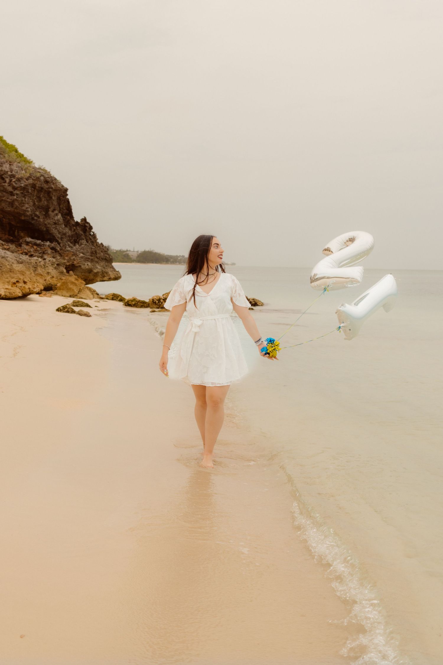 Dreamy beach scene with balloons, flowers, and figure in white dress.