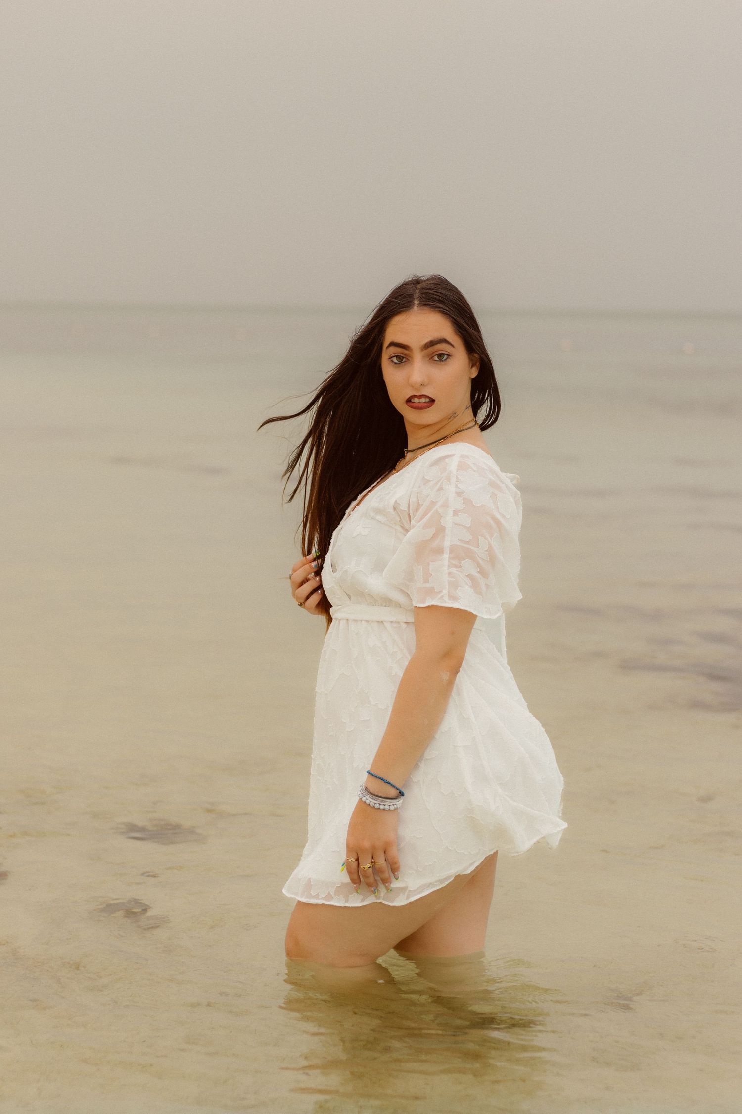 Model in white dress poses in shallow ocean water against moody sky at dusk.