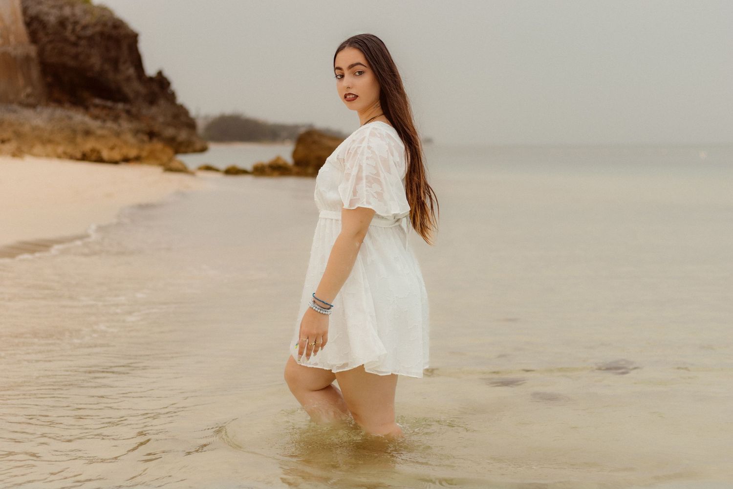 Model poses in white summer dress along serene beach shoreline.
