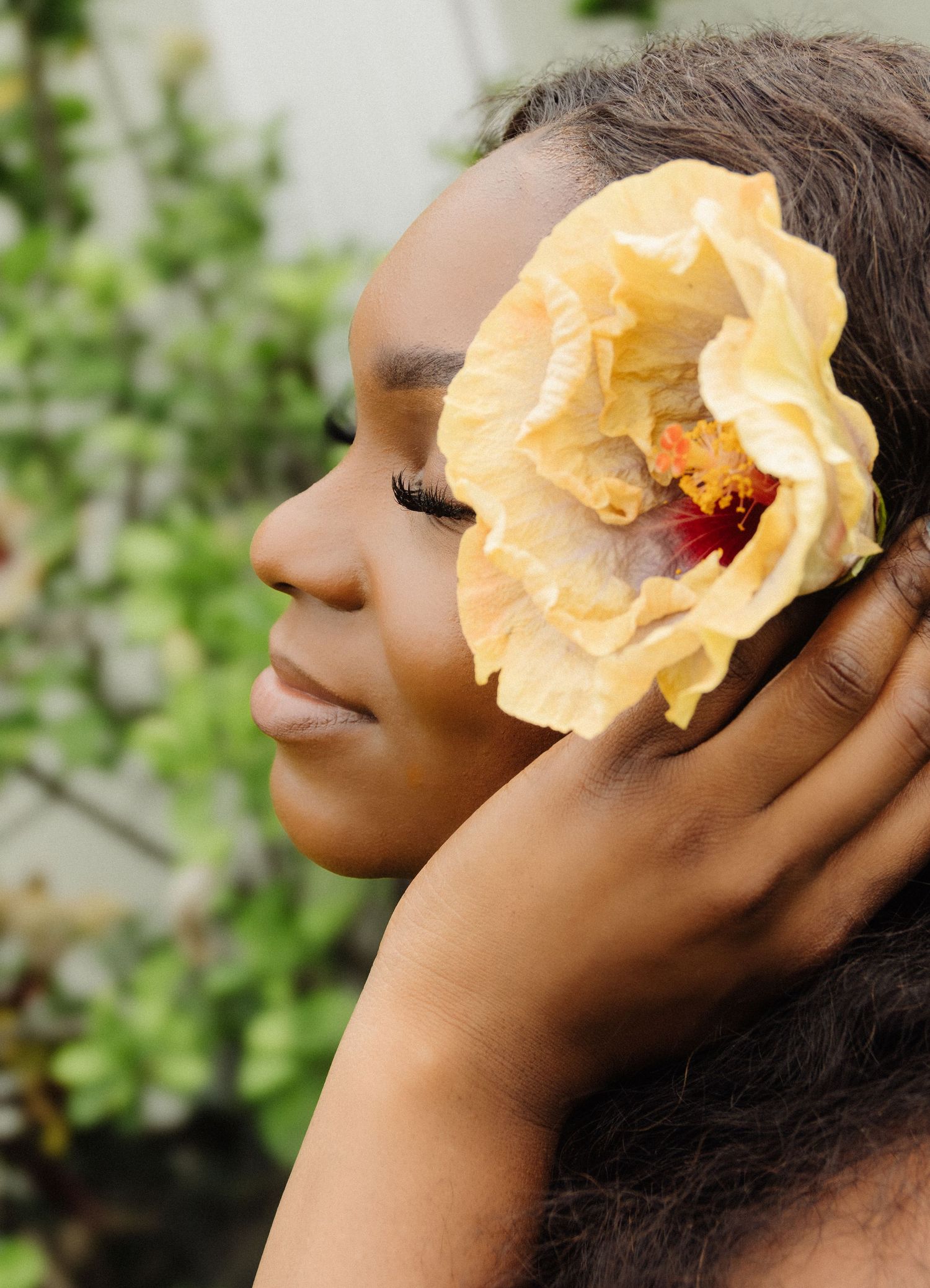 Person wearing yellow hibiscus flower in hair poses with eyes closed against green foliage.