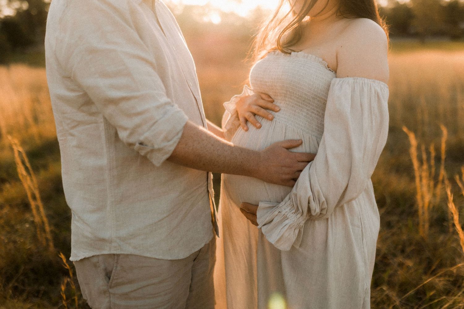 Intimate maternity photo of couple in white clothing standing in sunlit field.