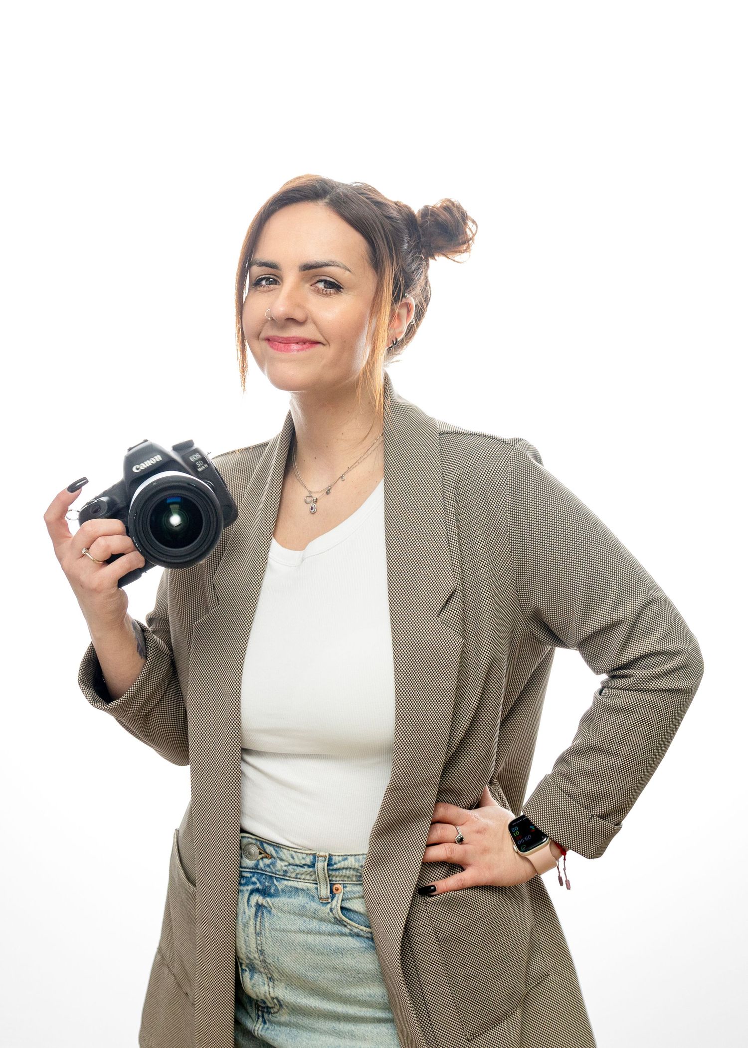 Photographer in beige blazer and white top posing with camera against white background.