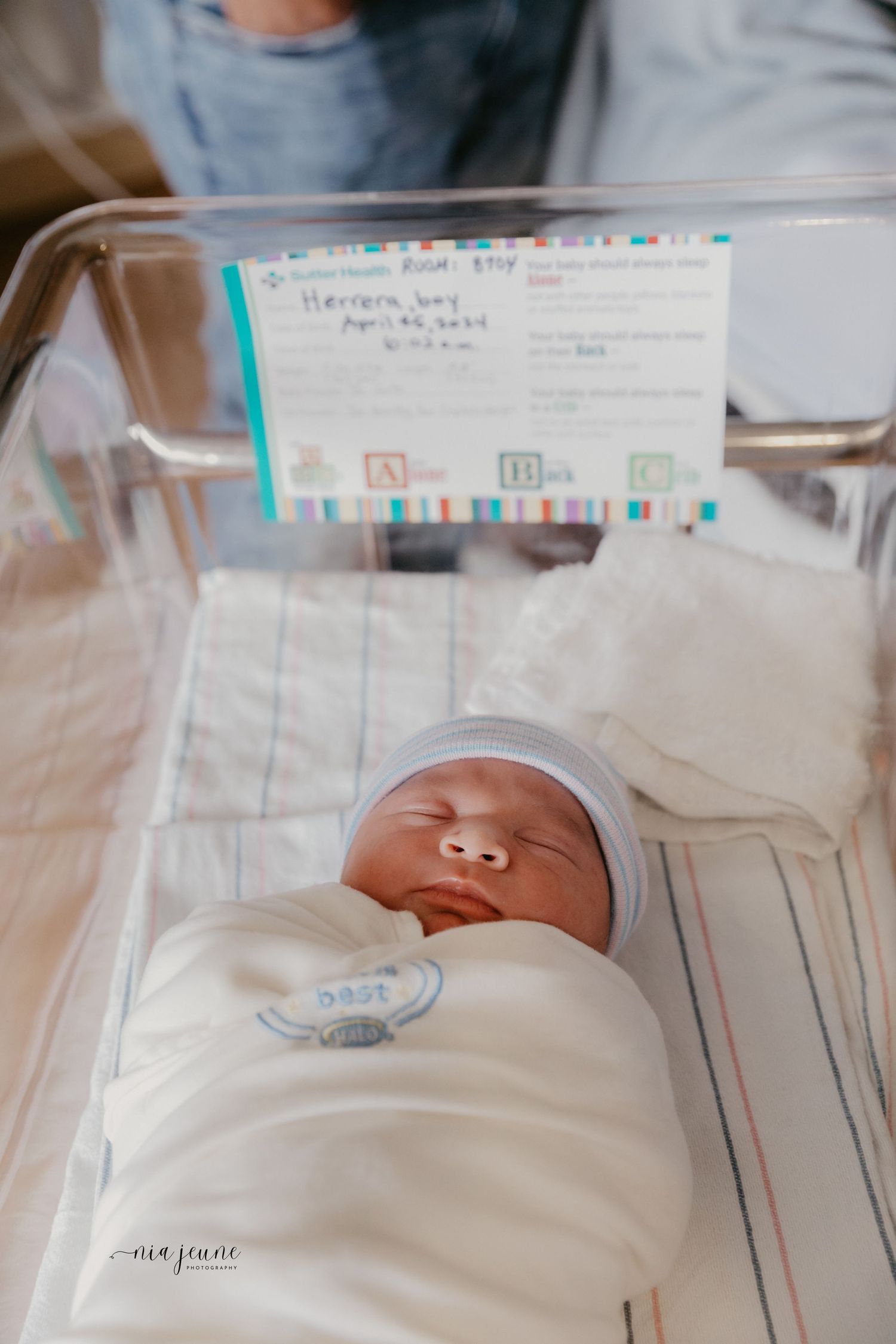 Newborn baby sleeping peacefully in hospital bassinet with medical information card visible.