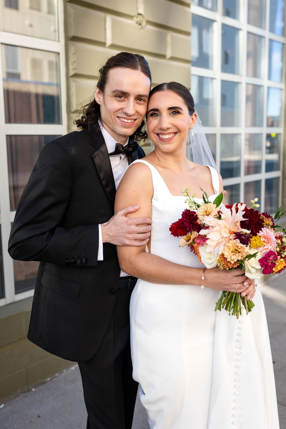 A newlywed couple embraces outside a building, with the bride holding a stunning burgundy and peach floral bouquet.