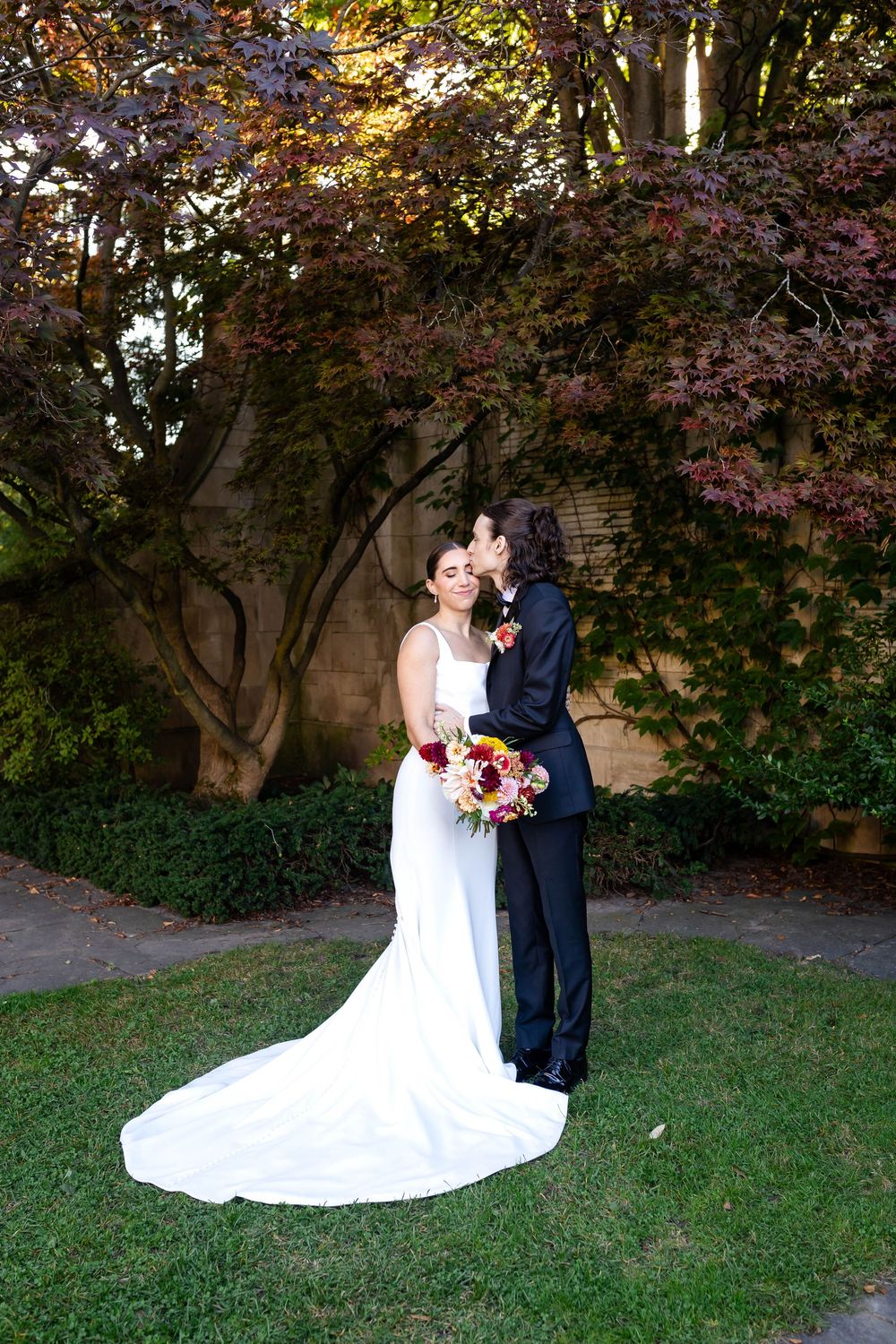 A wedding couple embraces under autumn maple trees with red foliage against a stone wall in a garden setting.