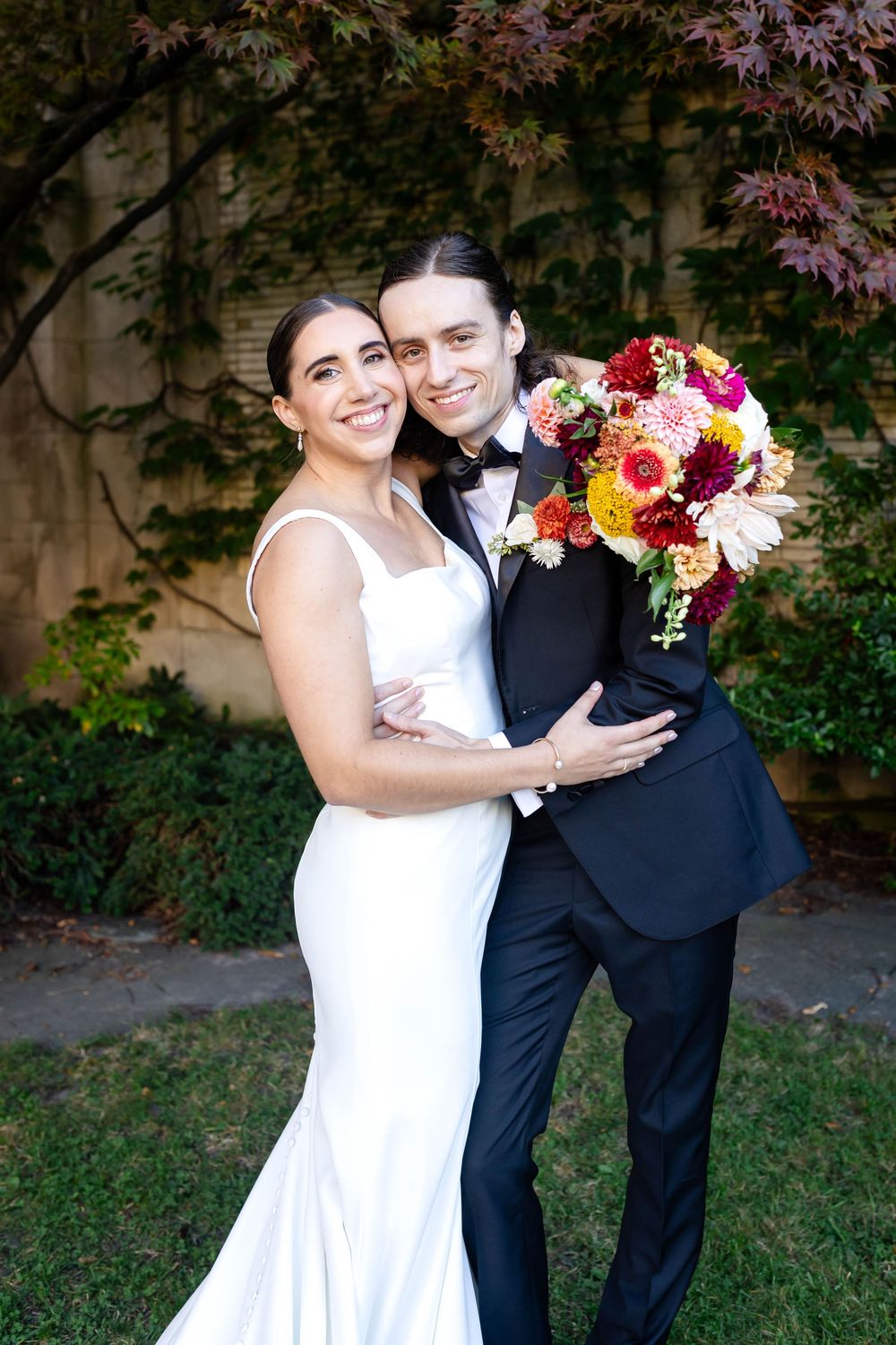 A wedding couple poses outdoors with vibrant pink and orange florals against a backdrop of Japanese maple trees.