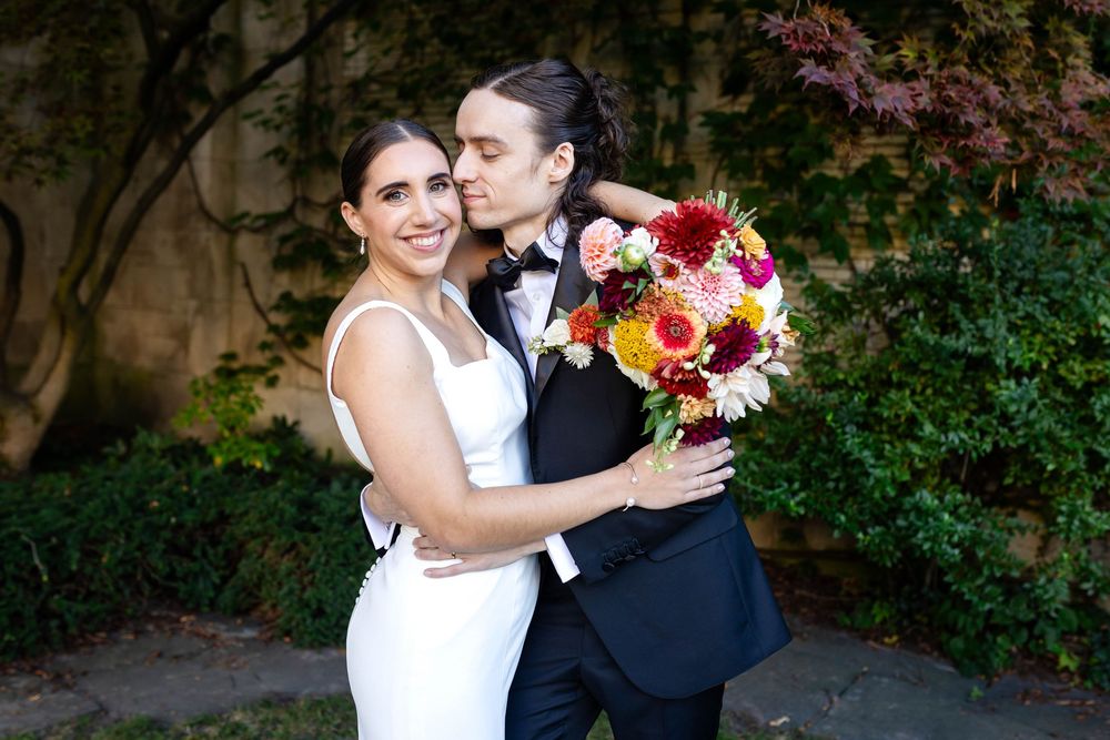 A joyful couple in elegant wedding attire embrace while holding a vibrant red and yellow floral bouquet in a garden setting.