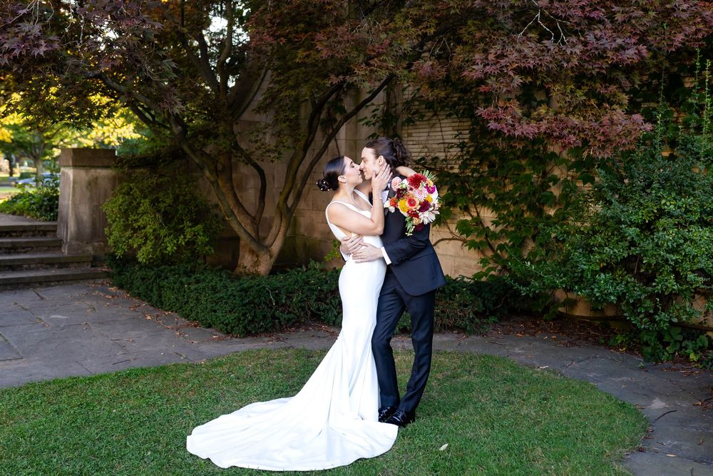 A couple in formal wedding attire embraces in a garden setting with flowering trees and greenery in the background.