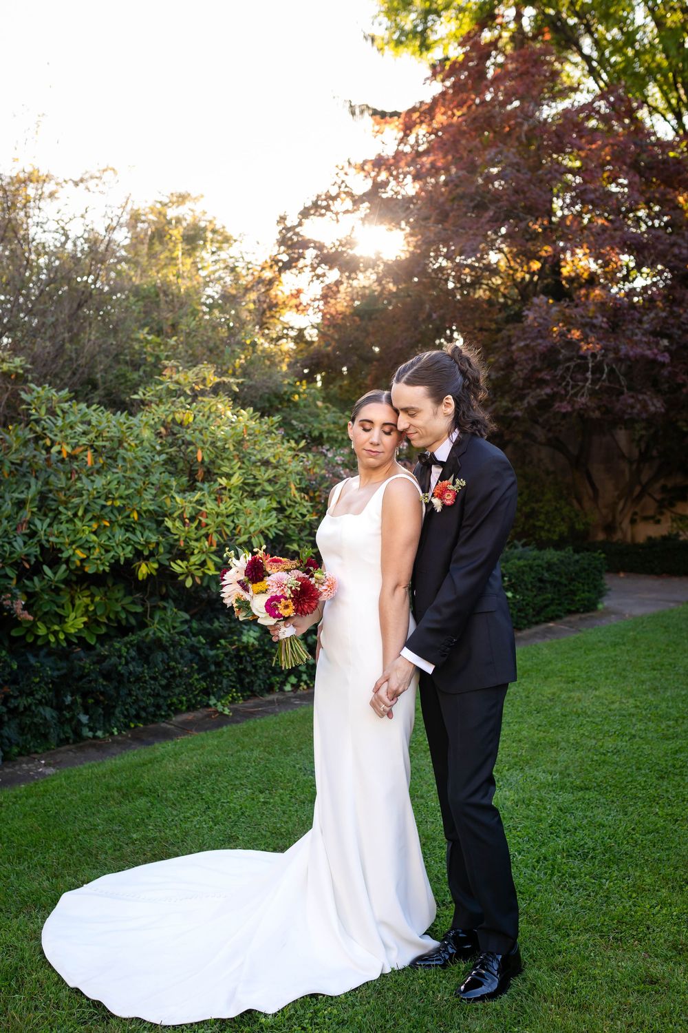 A couple poses together in formal wedding attire during golden hour in a garden setting with a vibrant bouquet.
