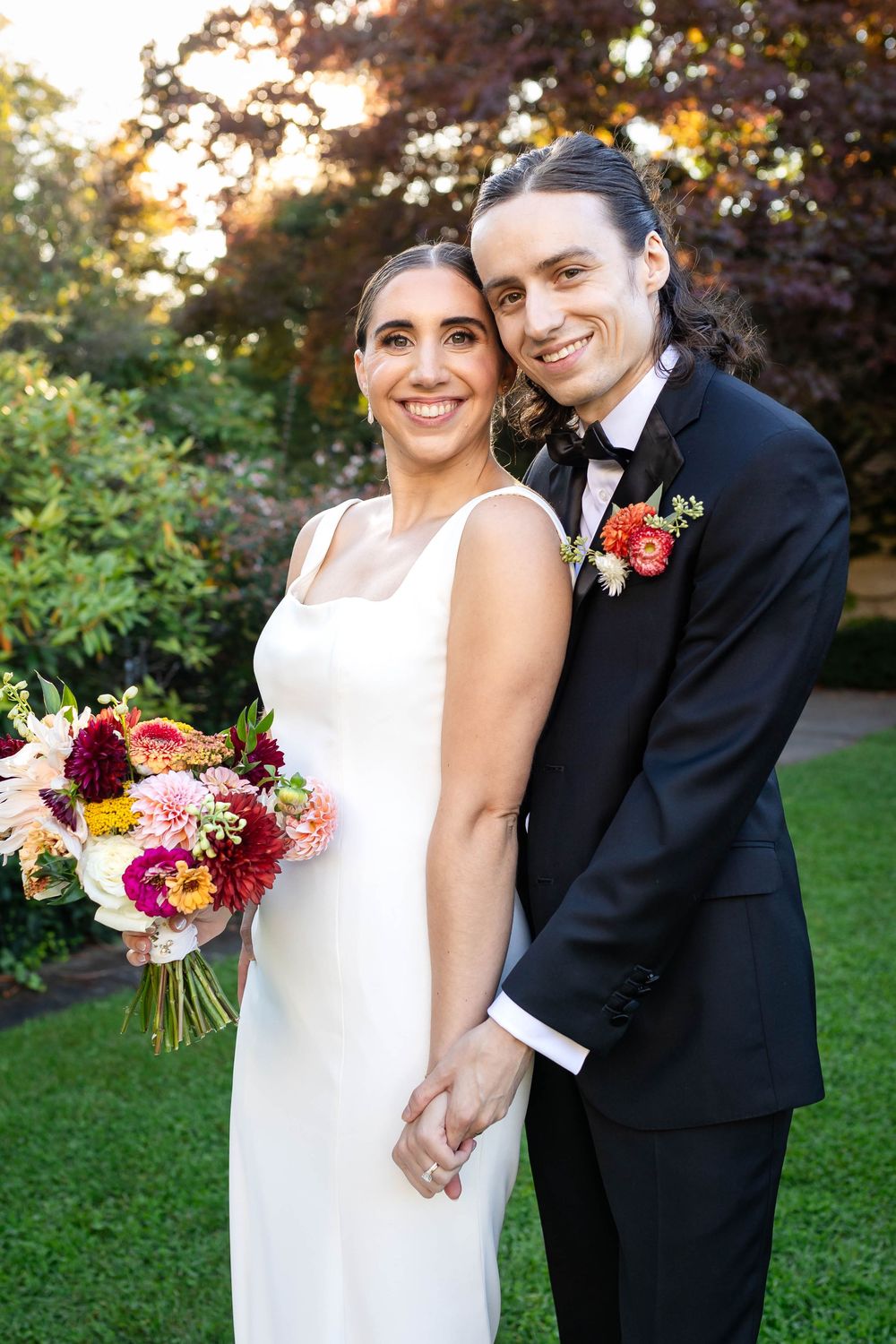 A couple stands together at their wedding celebration in a garden setting with a colorful autumn floral bouquet.