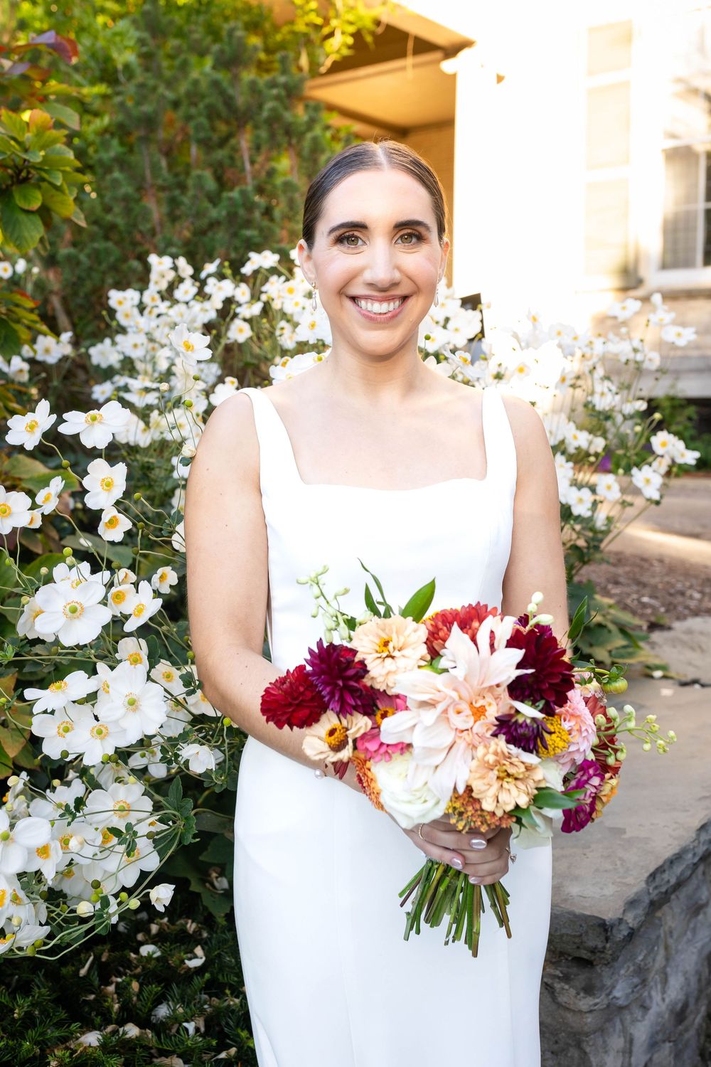 A smiling person in a white wedding dress holds a vibrant bouquet with burgundy and pink flowers against a backdrop of white blooms.