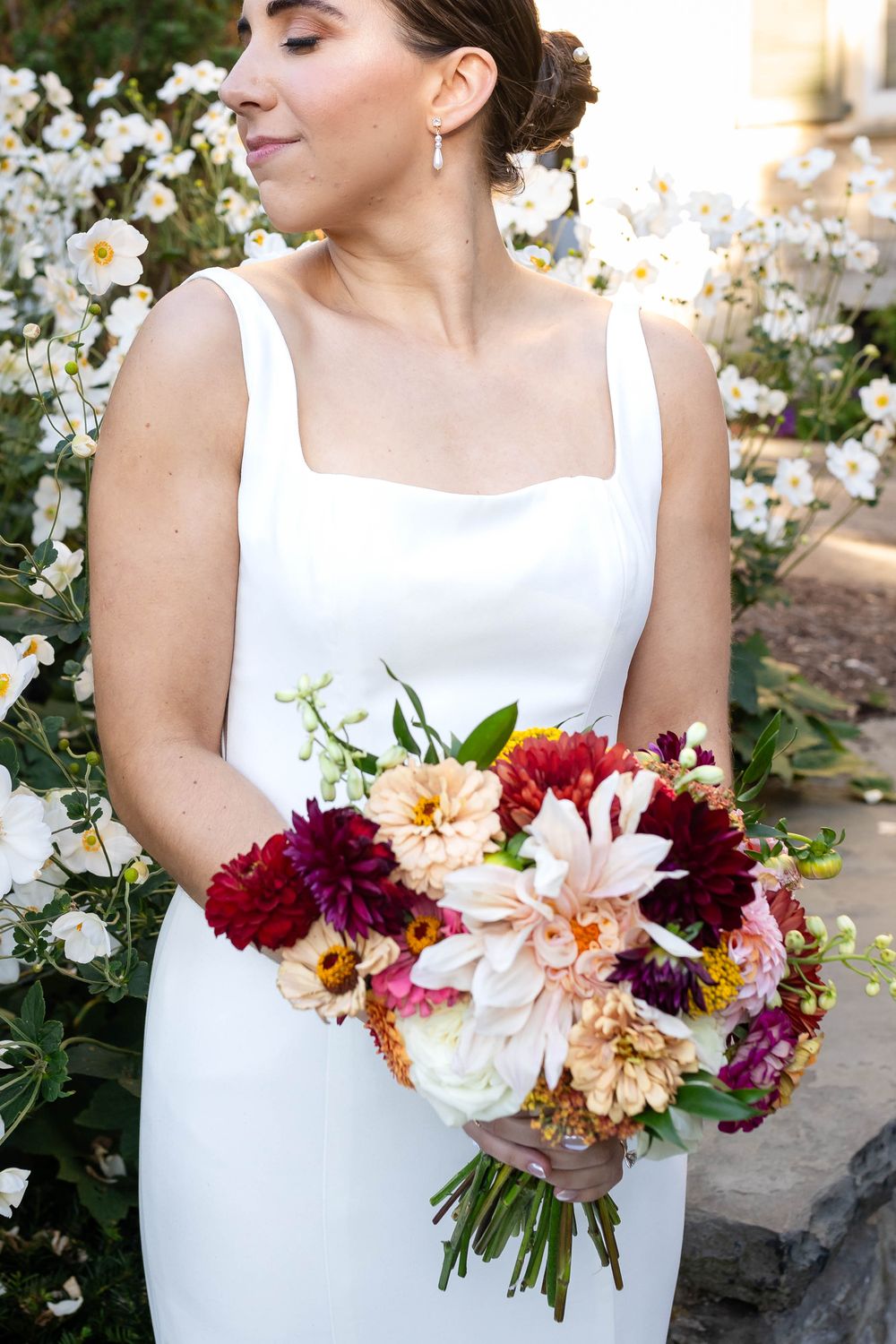 A bride in a sleek white dress holds a vibrant bouquet of dahlias, carnations and lilies against a garden backdrop.