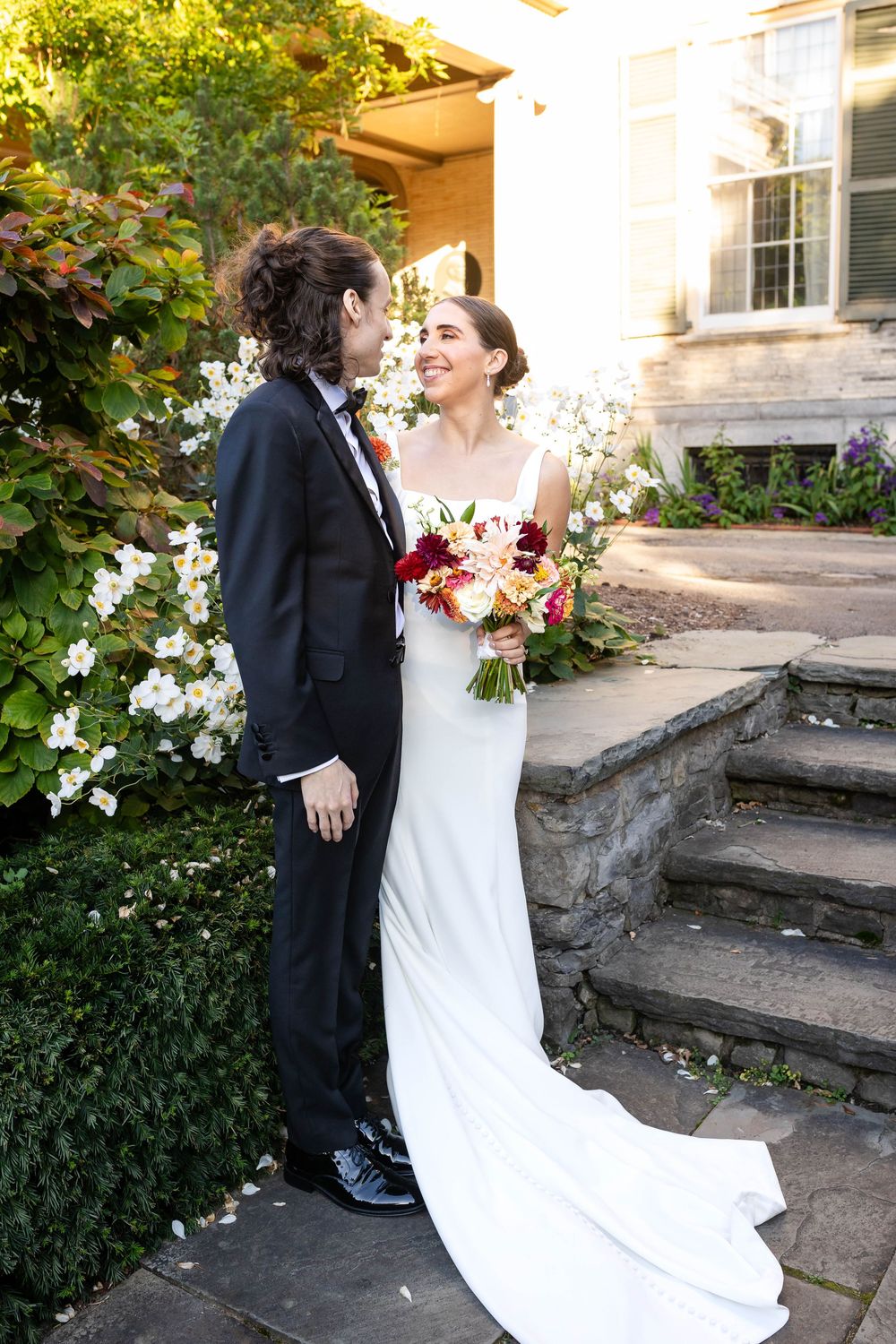 A couple in wedding attire share a romantic moment on stone steps surrounded by blooming flowers and greenery.