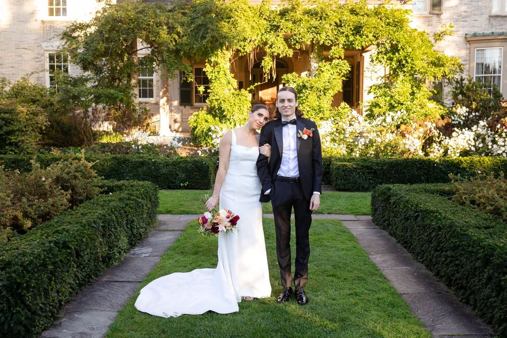 A wedding couple stands in a beautiful garden path lined with manicured hedges and blooming flowers on a sunny day.