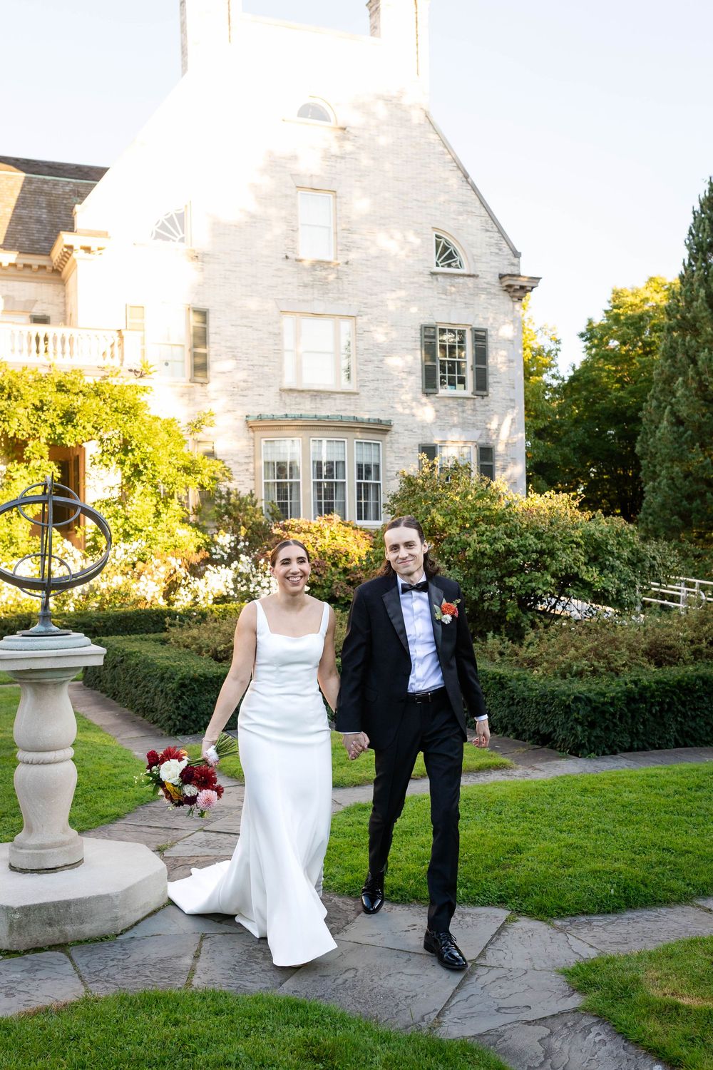 A couple stands on garden path beside sundial at elegant stone manor house with manicured landscaping.