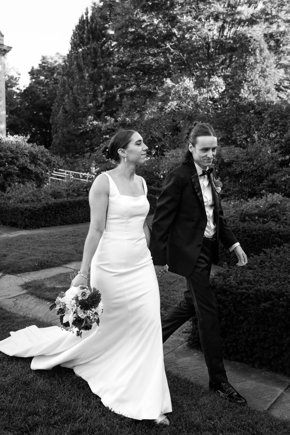 A black and white wedding photo showing a couple walking together in a garden, with the bride in a sleek white gown holding flowers.
