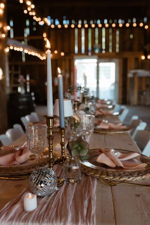 Long dining table with elegant place settings, taper candles, and string lights in a rustic barn setting.
