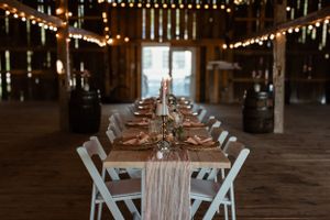 Long banquet table setup in a barn with white chairs and string lights creating an ambient atmosphere.